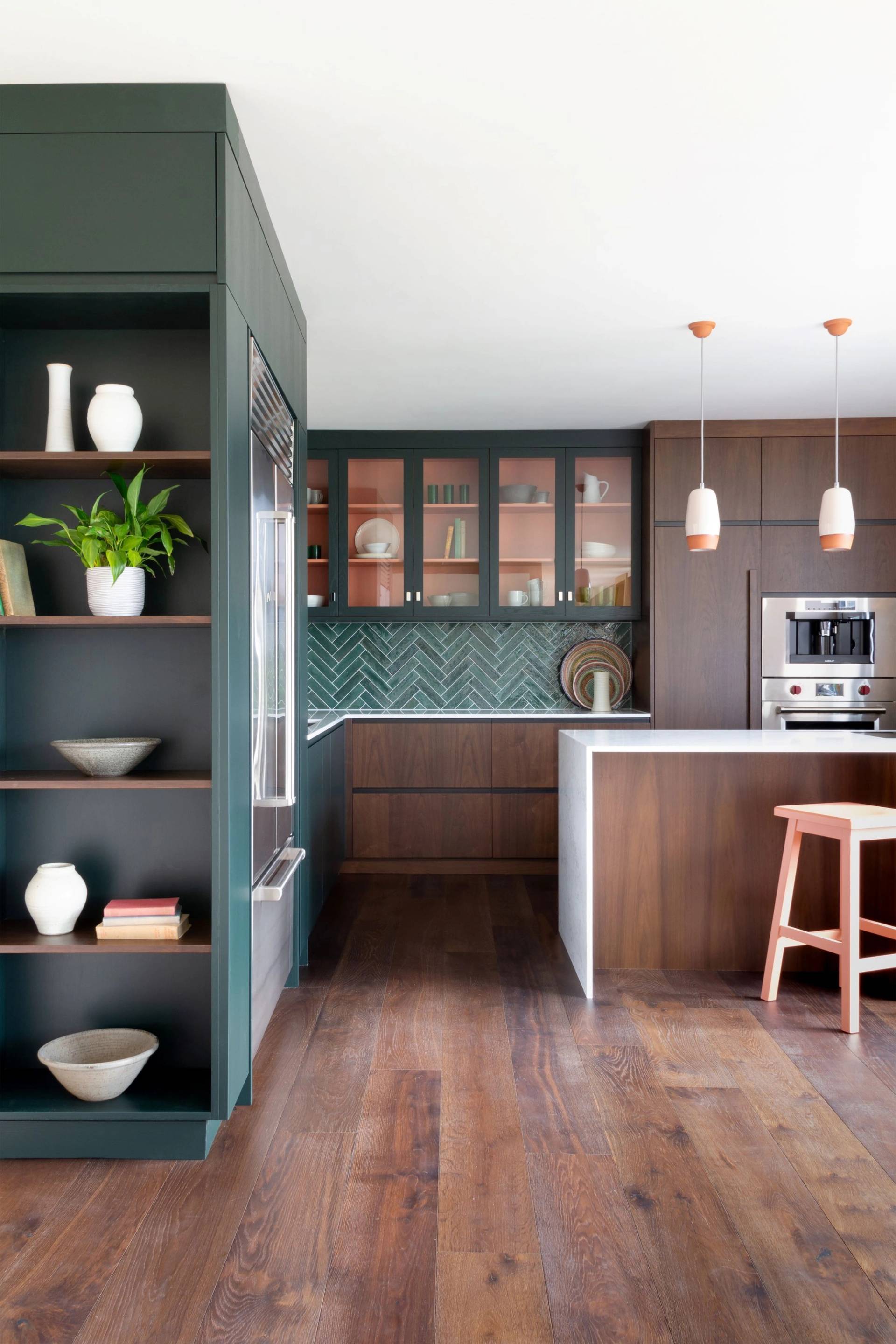A modern kitchen with dark wood flooring and cabinetry, green and white herringbone backsplash, and open shelving displaying plants and decor. A white countertop island has two pendant lights above and a wooden stool in front.