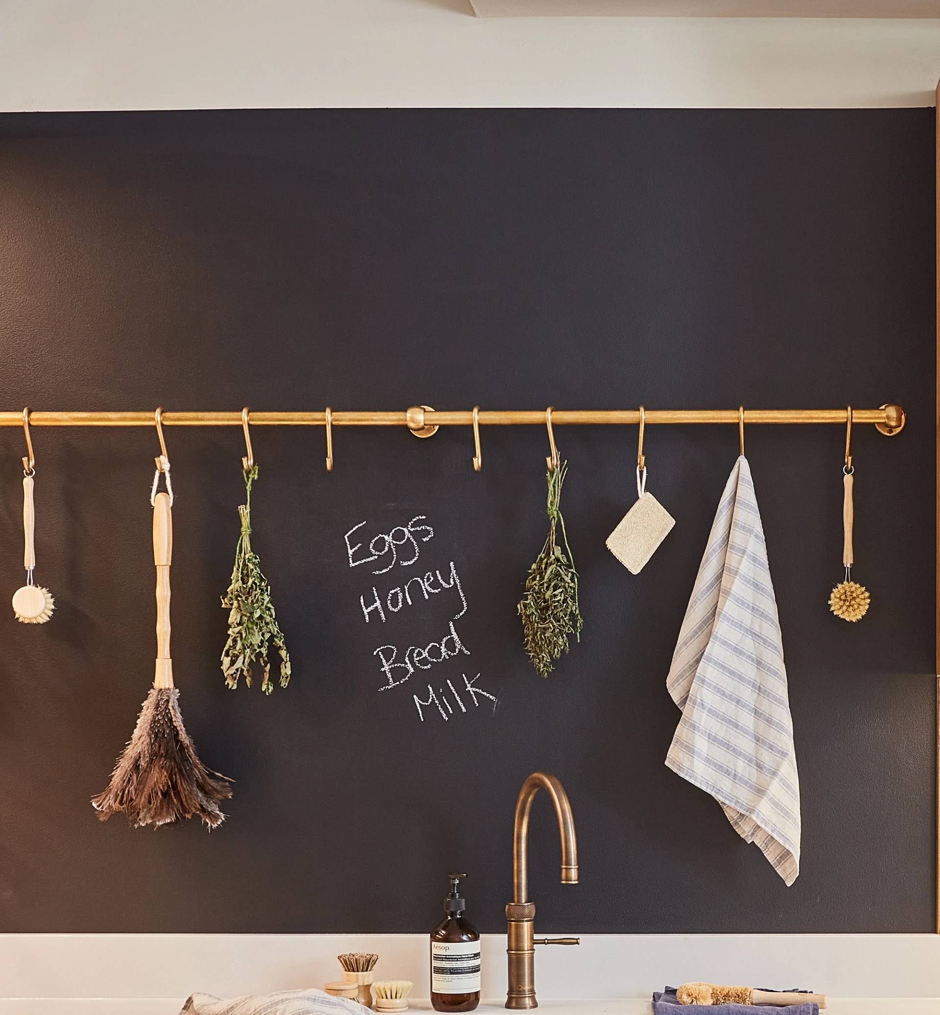 A kitchen wall with a dark chalkboard paint where 'eggs, honey, bread, milk' is written. A brass rod holds various hanging items, including herbs, towel, brush, and feather duster. Faucet soap dispenser are below.