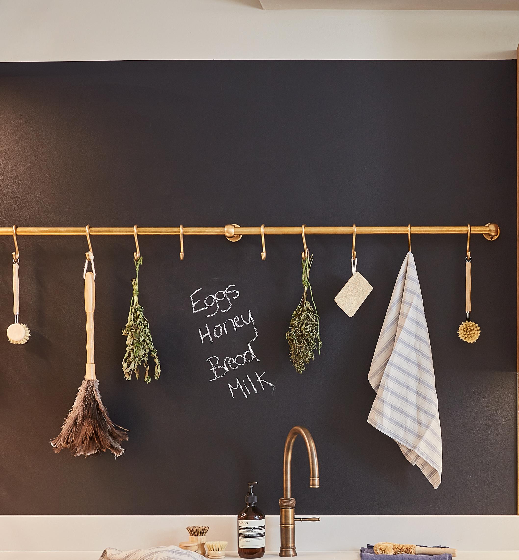 A kitchen wall with a dark chalkboard paint where 'eggs, honey, bread, milk' is written. A brass rod holds various hanging items, including herbs, towel, brush, and feather duster. Faucet soap dispenser are below.