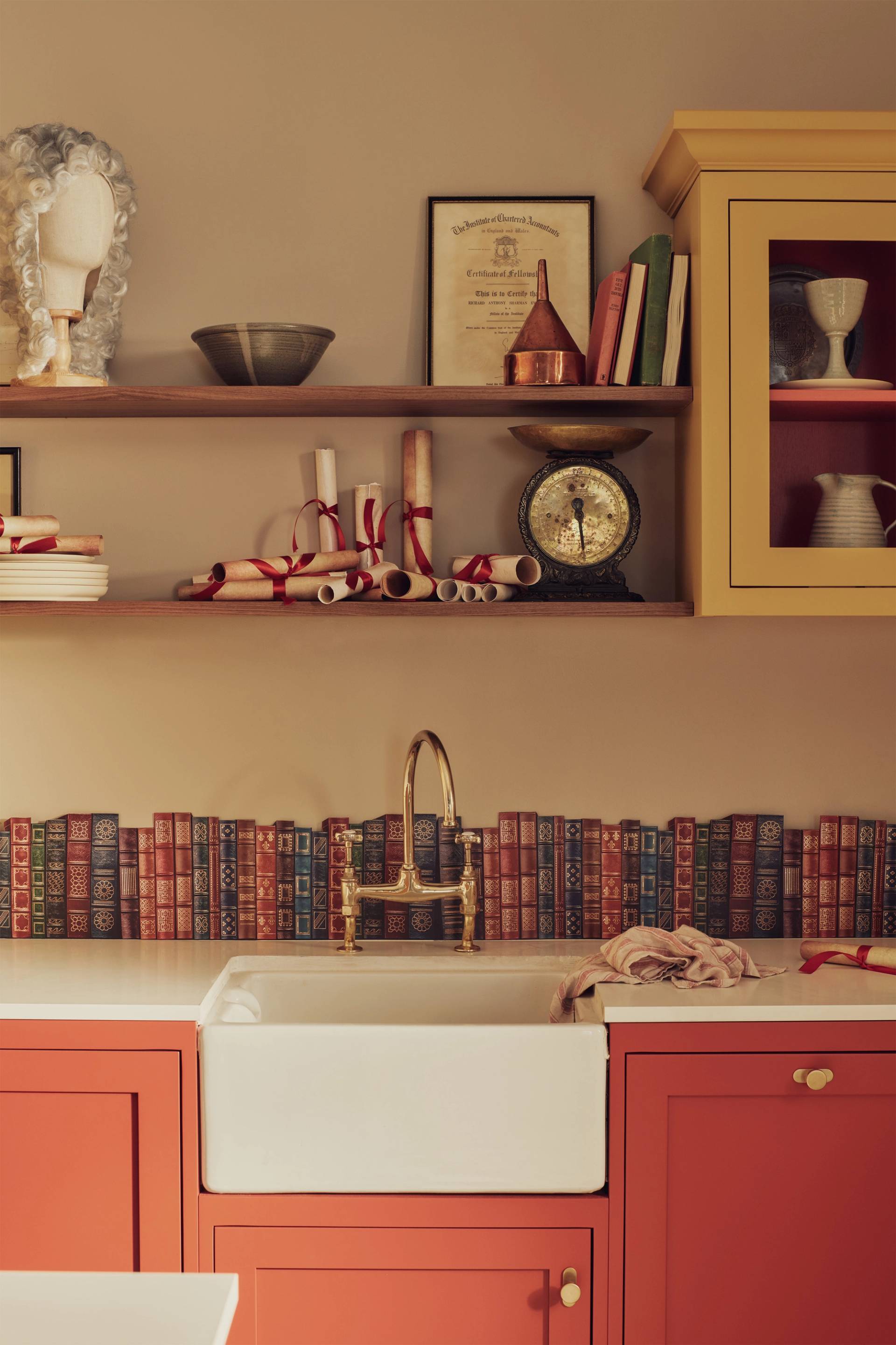 An eclectic kitchen scene featuring a white farmhouse sink with a gold bridge faucet, set against a unique backsplash made of antique book spines. The lower cabinets are a muted terracotta red, while open wooden shelves above display historical decor: a powdered wig on a stand, rolled scrolls tied with red ribbons, a vintage weighing scale, and a framed certificate. A mustard yellow cabinet adds a pop of color to the warm, academic-inspired space.