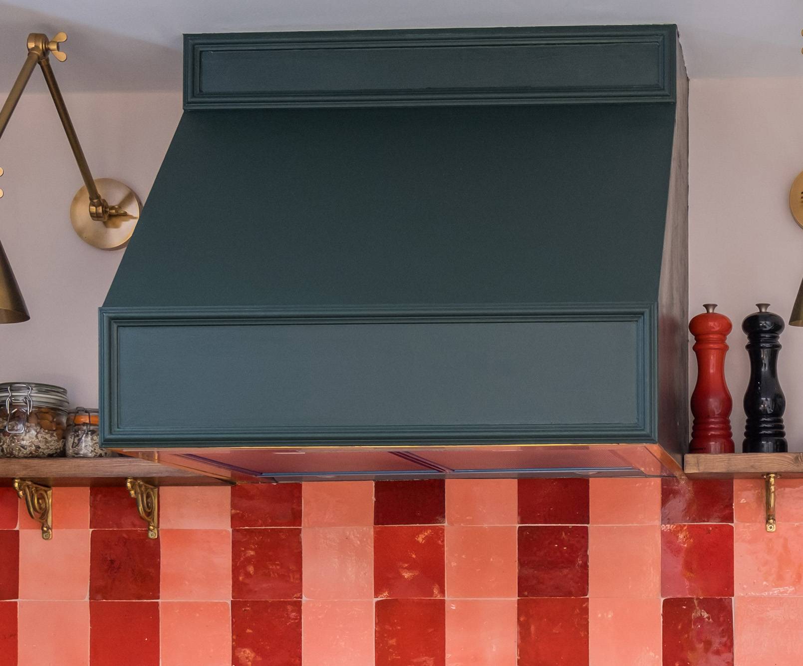 A dark green kitchen range hood above a shelf holding spice jars, a black pepper grinder, and a red salt grinder, with a red and pink checkered tile backsplash and decorative wall lights.