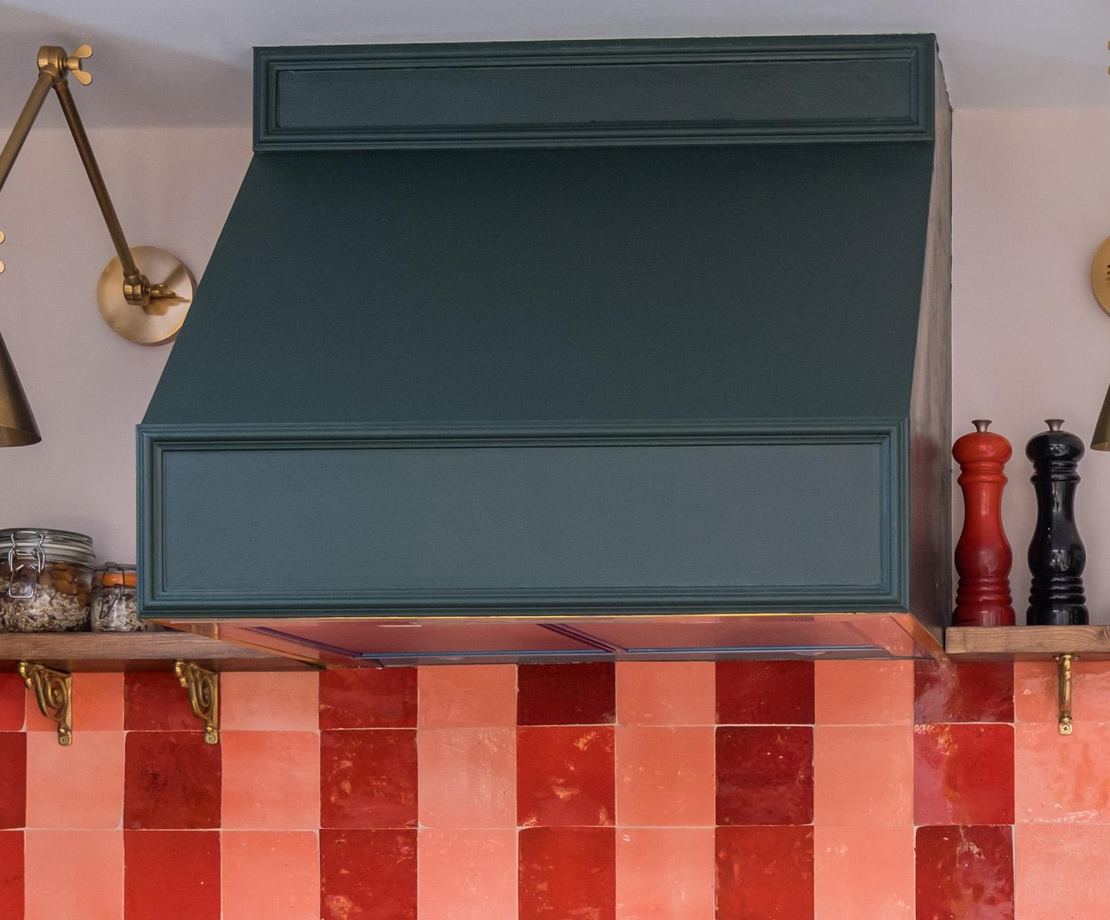 A dark green kitchen range hood above a shelf holding spice jars, a black pepper grinder, and a red salt grinder, with a red and pink checkered tile backsplash and decorative wall lights.