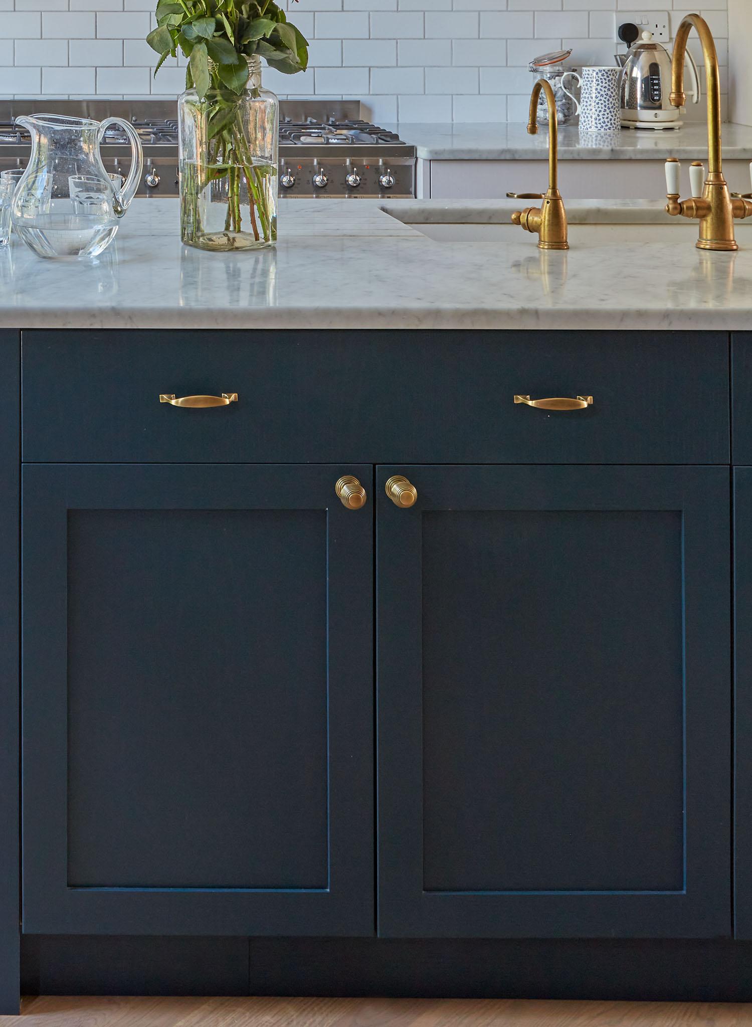 A kitchen island with dark blue cabinets, gold handles, a white marble countertop, gold faucet fixtures, a glass pitcher, a vase with green foliage, and a stove with a subway tile backsplash in the background.