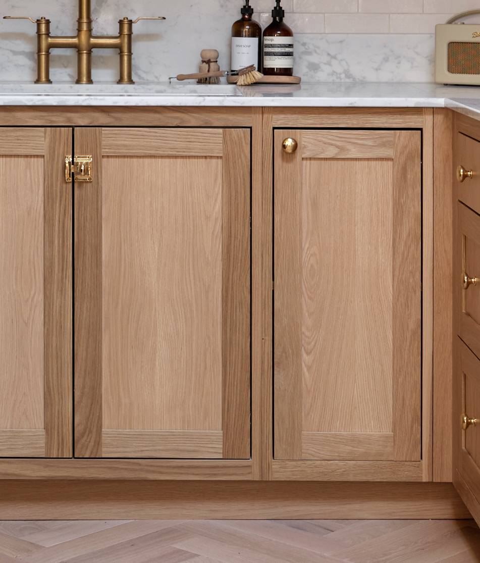 Light wood kitchen cabinets with gold hardware, a white marble countertop, and a brass faucet. Bottles, a brush, and a small radio sit on the counter. The backsplash is white subway tile.