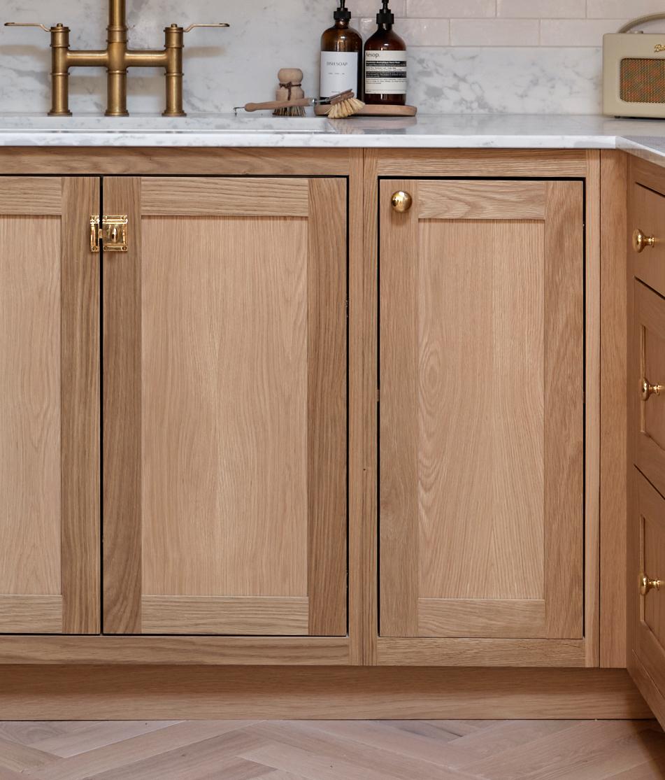 Light wood kitchen cabinets with gold hardware, a white marble countertop, and a brass faucet. Bottles, a brush, and a small radio sit on the counter. The backsplash is white subway tile.