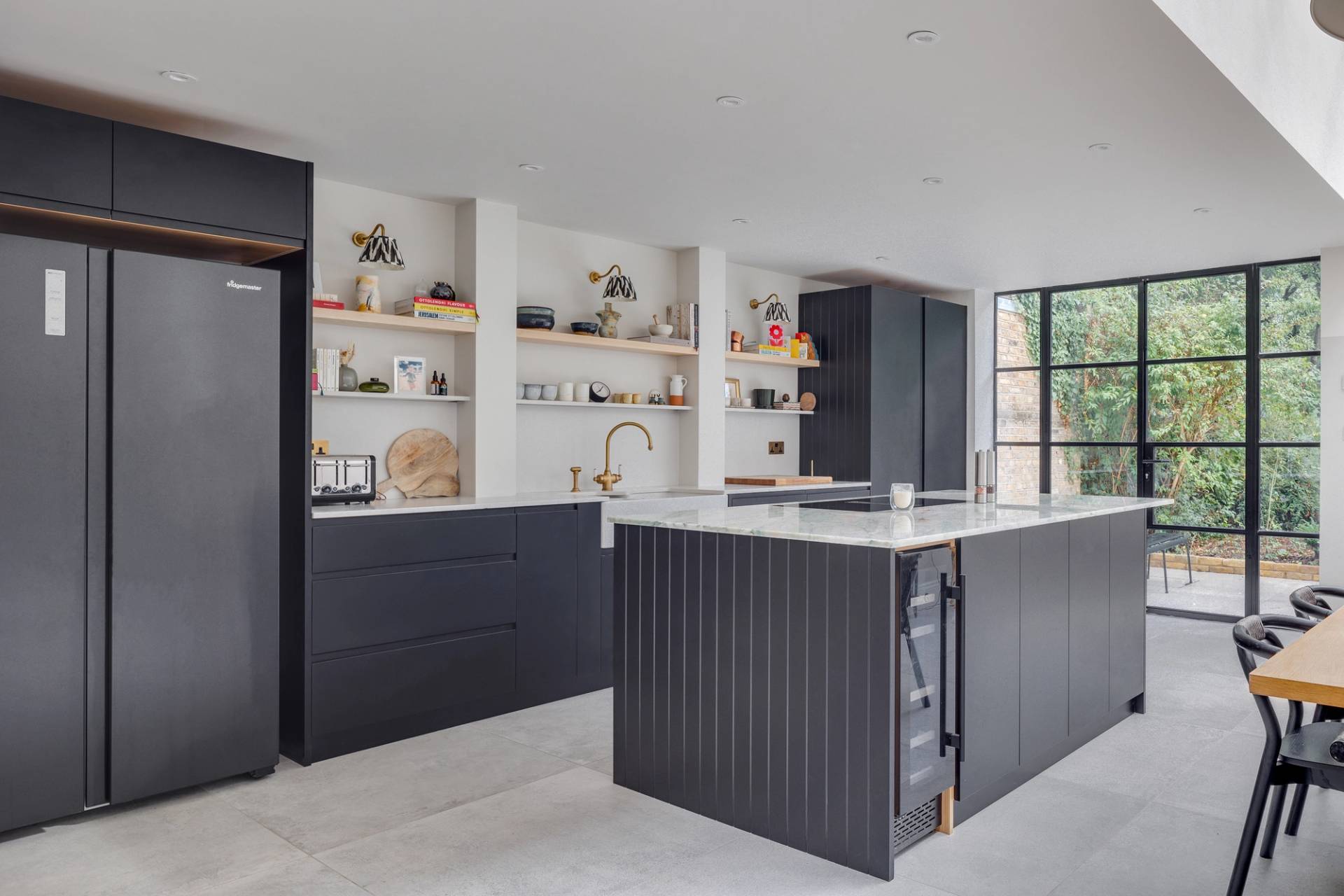 Modern kitchen with a sleek island, dark cabinetry, open shelves displaying kitchenware, and a large refrigerator. A wall of windows offers natural light and a view of greenery. Neutral tones and minimalist design create a contemporary atmosphere.
