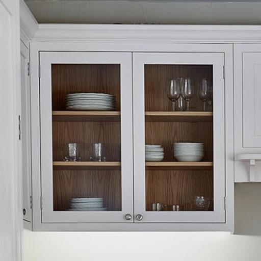 A white kitchen cabinet with glass doors displays neatly stacked white plates, bowls, and various glassware on three wooden shelves. The cabinet is built into a white kitchen wall.