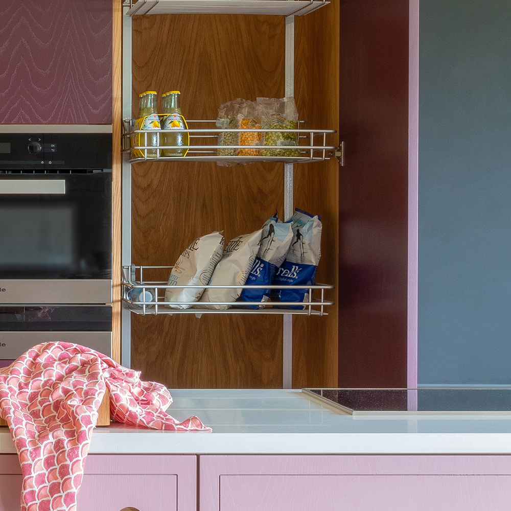 An open kitchen pantry with shelves holding bottled drinks, bags of snacks, and food containers. A pink and white dish towel is draped over a pink countertop in the foreground.