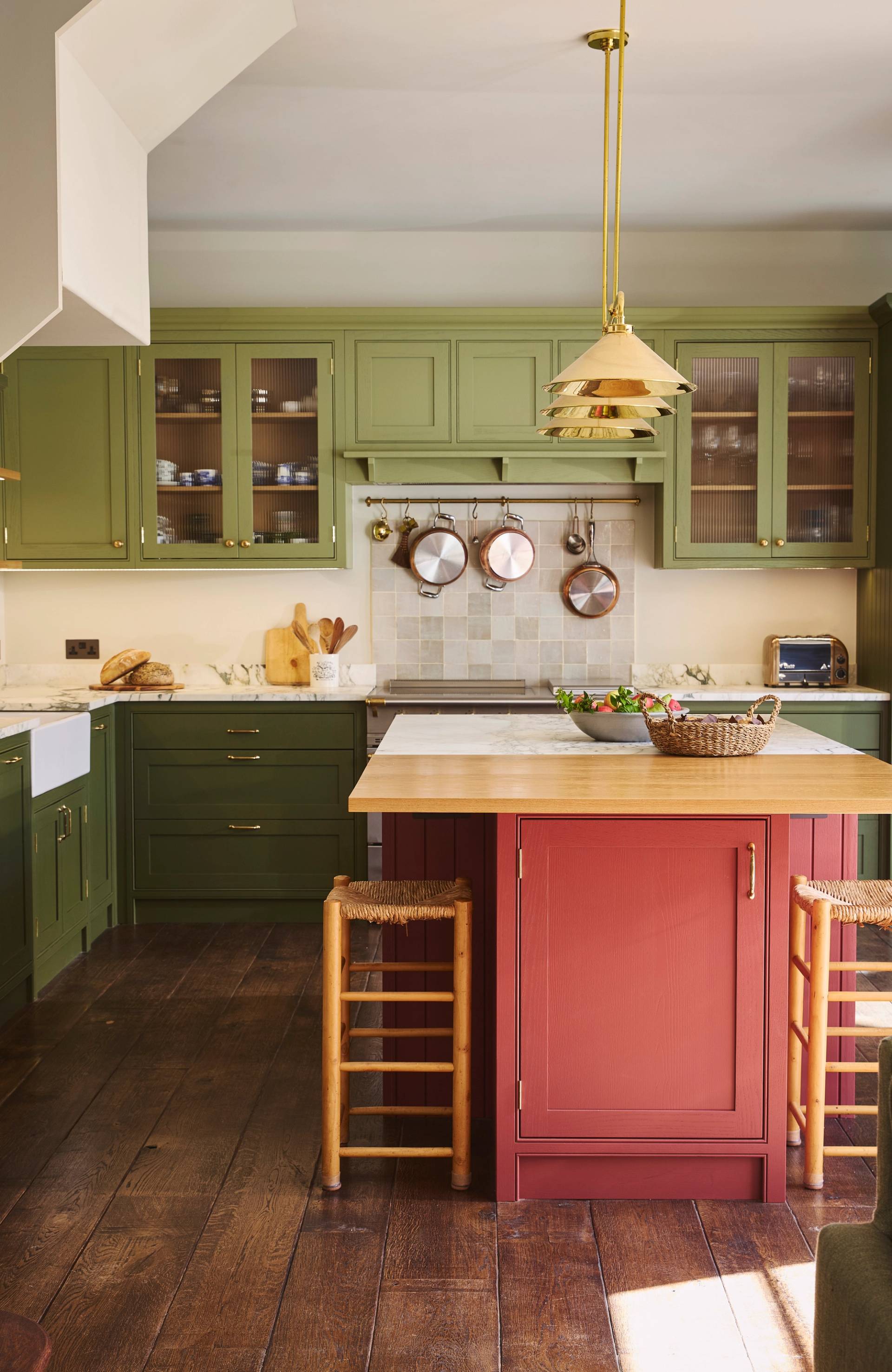 A modern kitchen with olive-green cabinets, open shelving with books and baskets, a small folding bicycle stored under a desk, and sunlight streaming through a glass door. A framed poster and potted plant are on the desk.