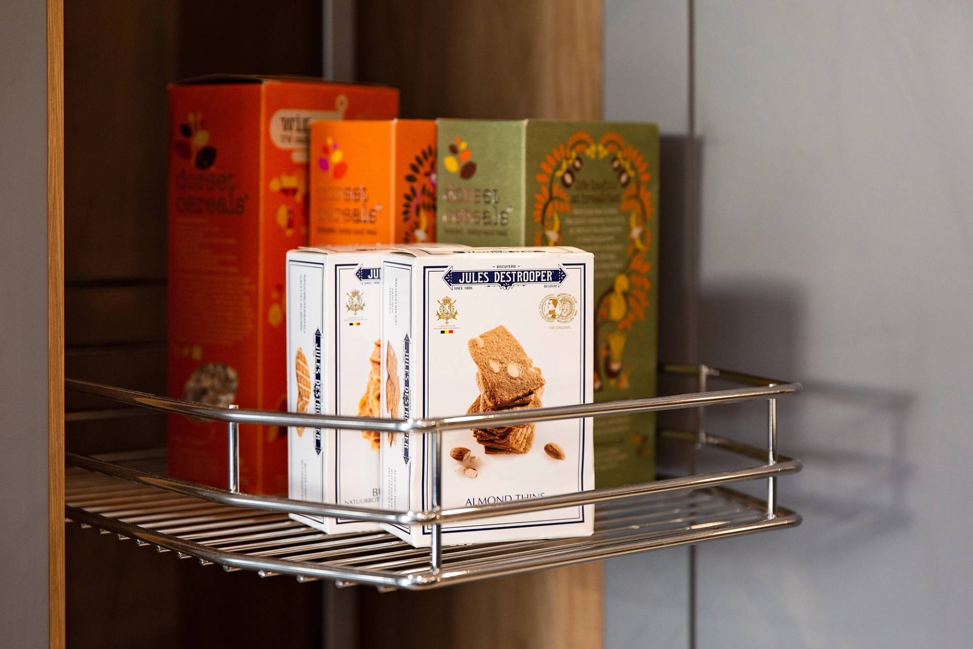 A shelf inside a cabinet holds various boxes of snacks, including Jules Destrooper almond thins and other colorful boxes of crackers or cookies. The snack boxes are neatly arranged on a metal wire shelf.