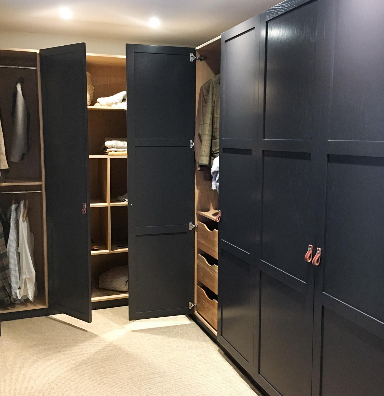 A modern wardrobe with dark paneled doors, some open to reveal hanging clothes, shelves with folded items, and drawers, set in a carpeted room with overhead lighting.