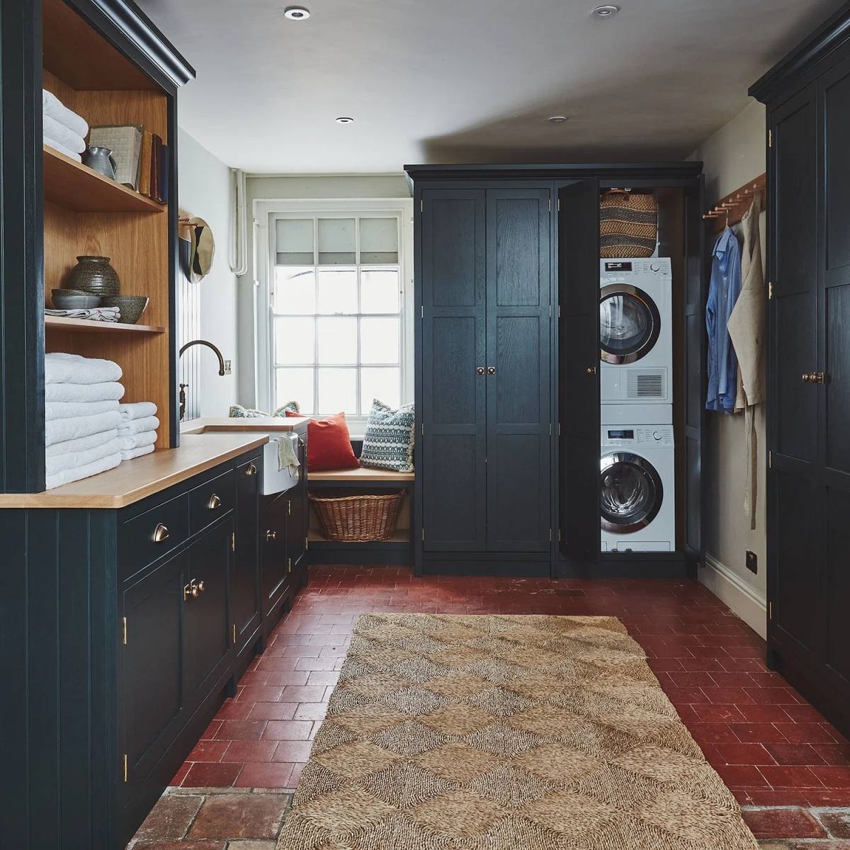 Laundry room with dark cabinets, a stacked washer and dryer, brick floor, and a woven rug. Wooden countertop holds folded towels. A window seat with cushions is in the corner, and there are hangers with shirts on the right.