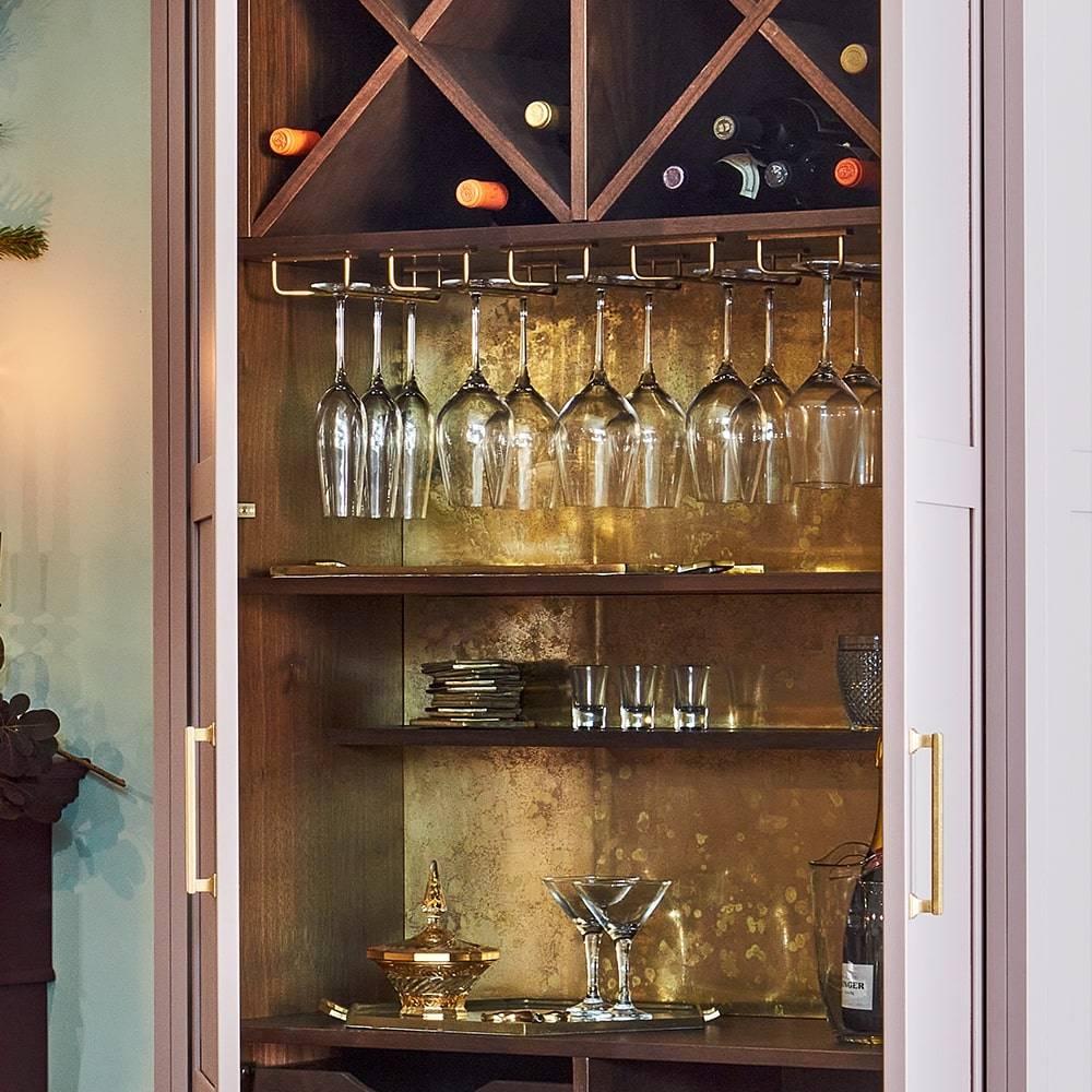 A home bar cabinet with wine bottles, hanging wine glasses, shelves with glassware, and gold-colored decor against a gold-toned interior. The cabinet doors are open, revealing the organized bar setup.