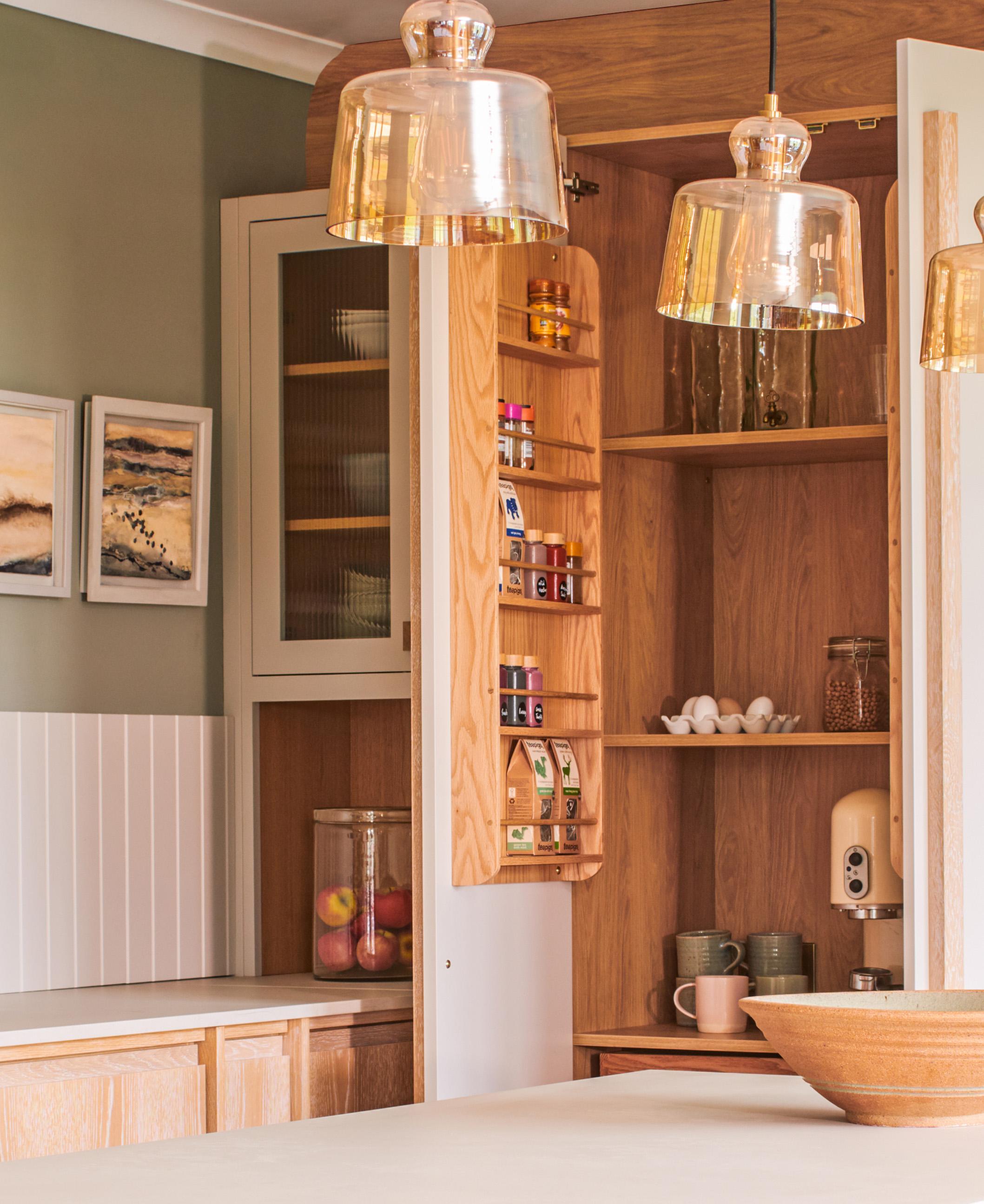 A cozy kitchen corner features wooden cabinets with an open pantry displaying spices and jars. Two pendant lights hang above a kitchen island. Decorative framed art adorns the green wall.
