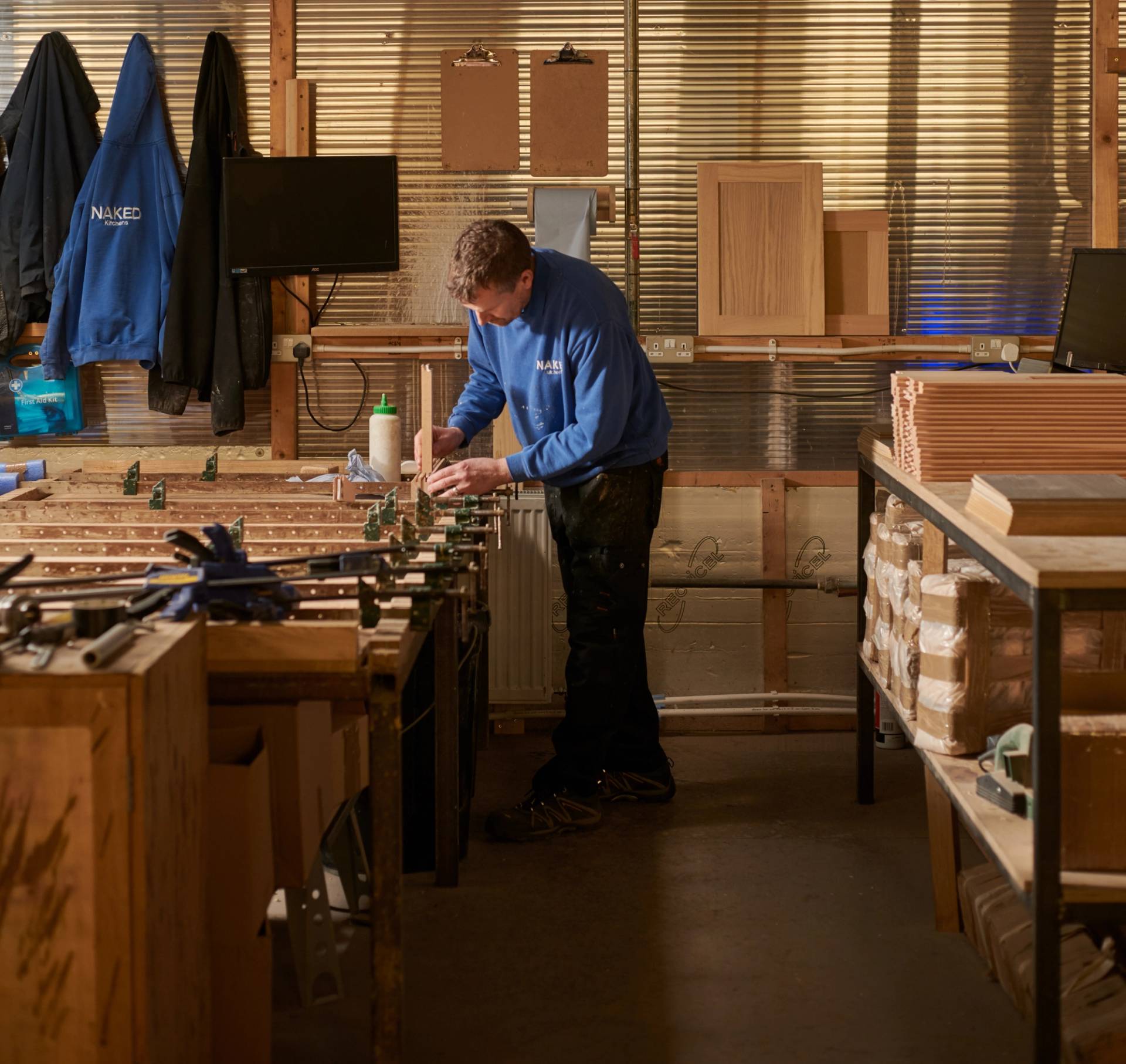 A person in a blue shirt works in a woodshop, using tools on a large table. The space has wooden cabinets, shelving with boxes, and clipboards on the wall. Two jackets hang nearby, and a monitor is visible in the background.