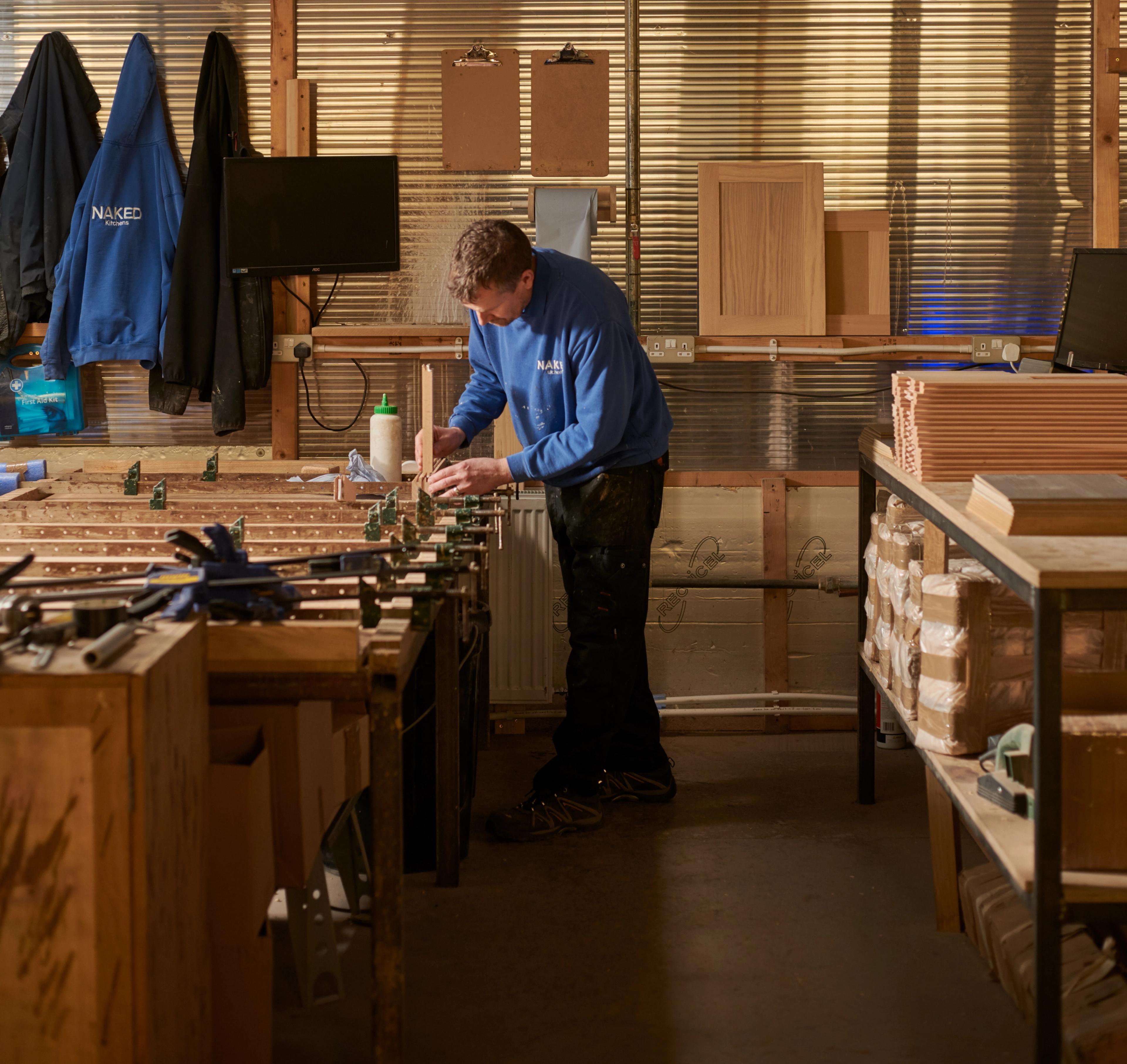 A person in a blue shirt works in a woodshop, using tools on a large table. The space has wooden cabinets, shelving with boxes, and clipboards on the wall. Two jackets hang nearby, and a monitor is visible in the background.