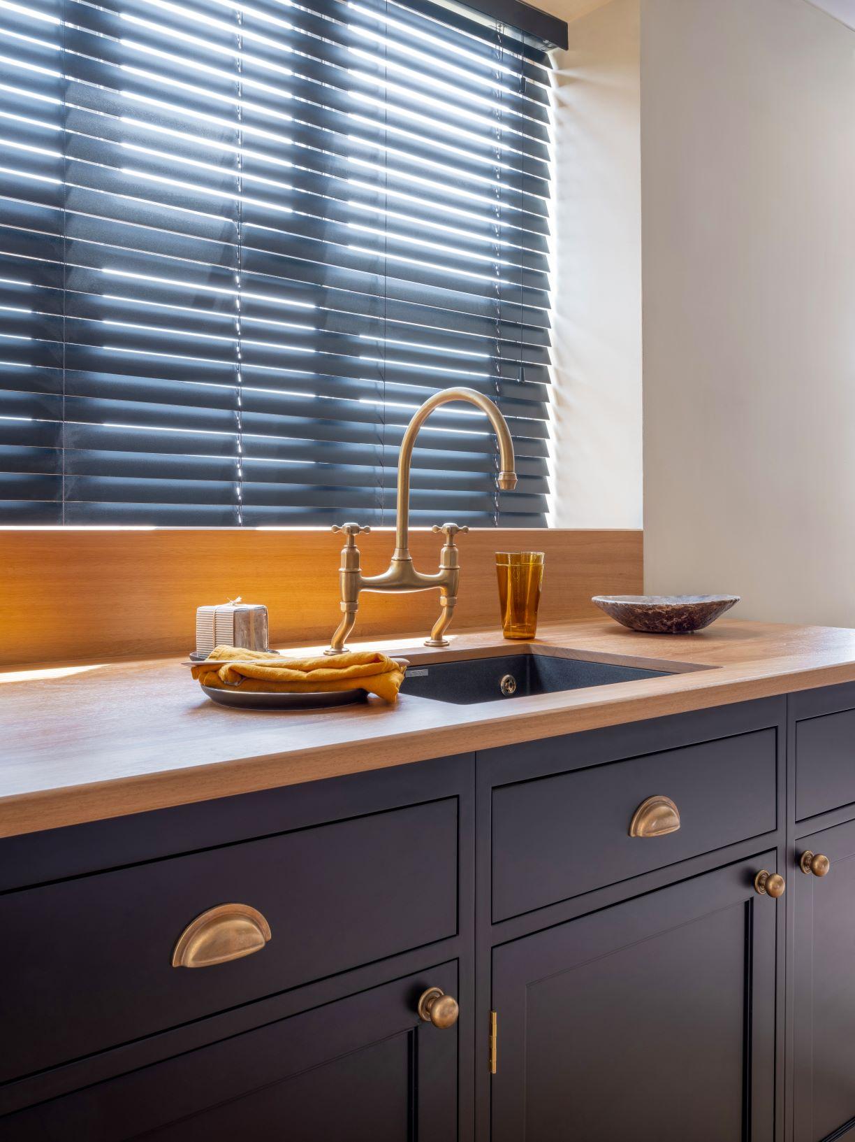 A modern kitchen features dark blue cabinets and a wooden countertop. Brass fixtures complement the faucet and cabinet handles. A bowl and glass sit near the sink, with a towel folded nearby. Horizontal blinds cover the window in the background.
