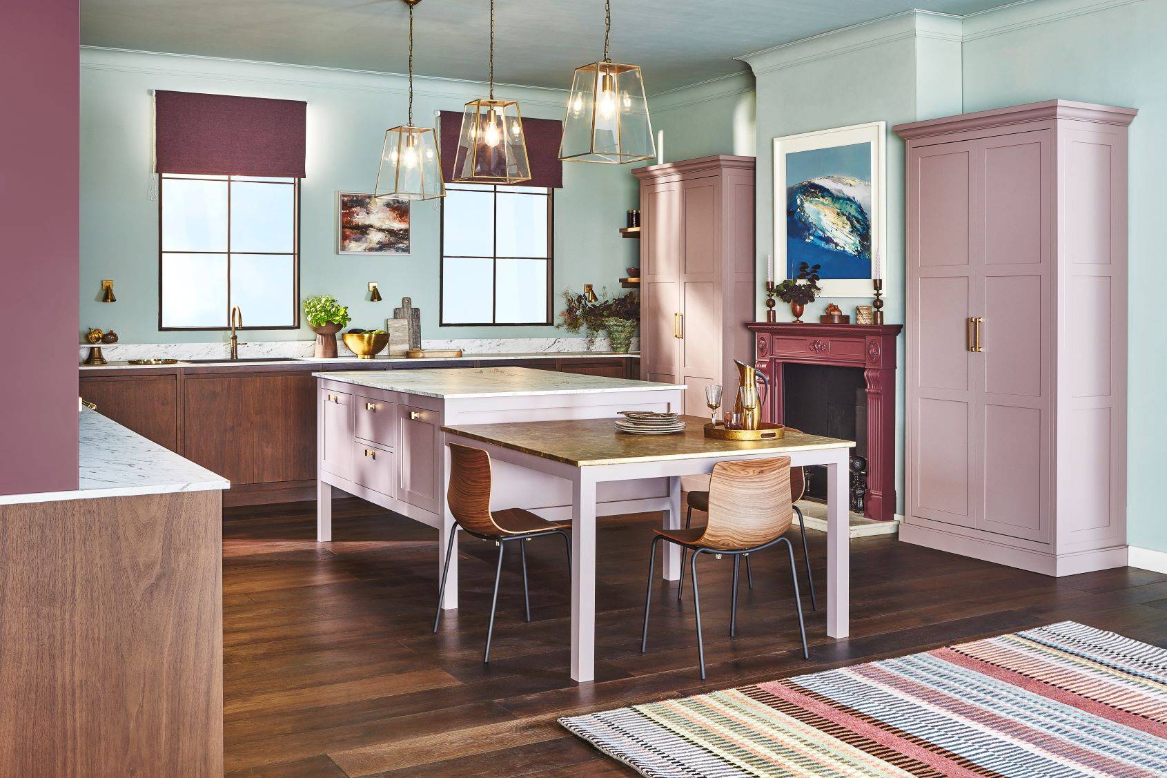 A modern kitchen with a pastel color scheme features wooden flooring, a central island with two chairs, and pendant lights. Cabinets are a soft mauve, and there is a rug near the island. Artwork and plants decorate the space, with windows providing natural light.