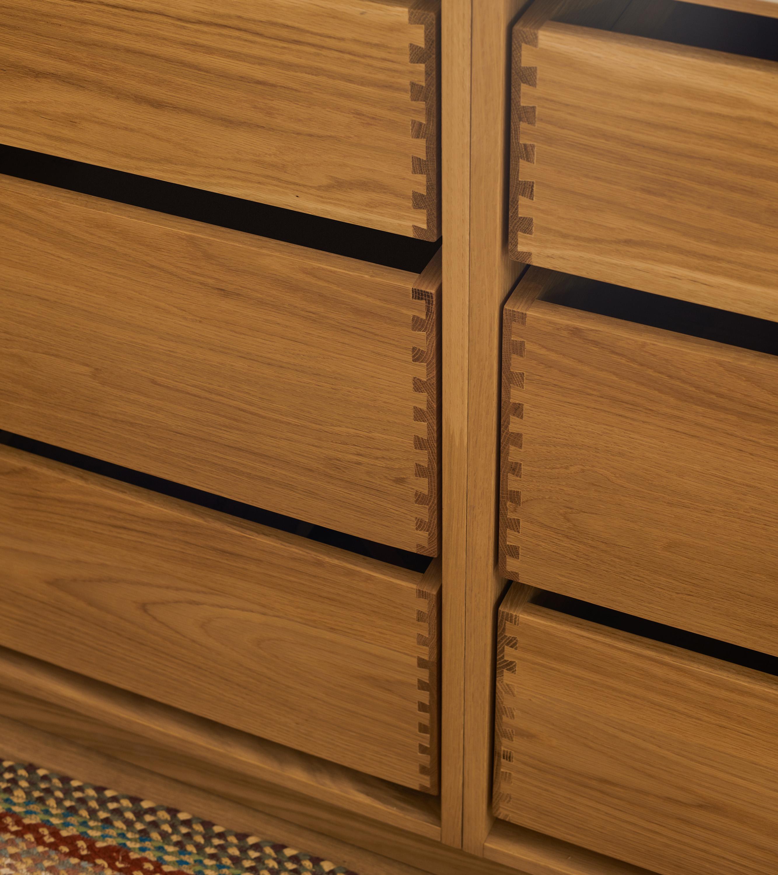 Close-up of a wooden kitchen cabinet with six visible drawers featuring dovetail joints. The cabinet has a natural wood finish and is topped with a light-colored countertop. A dark pot is partially visible on the counter.
