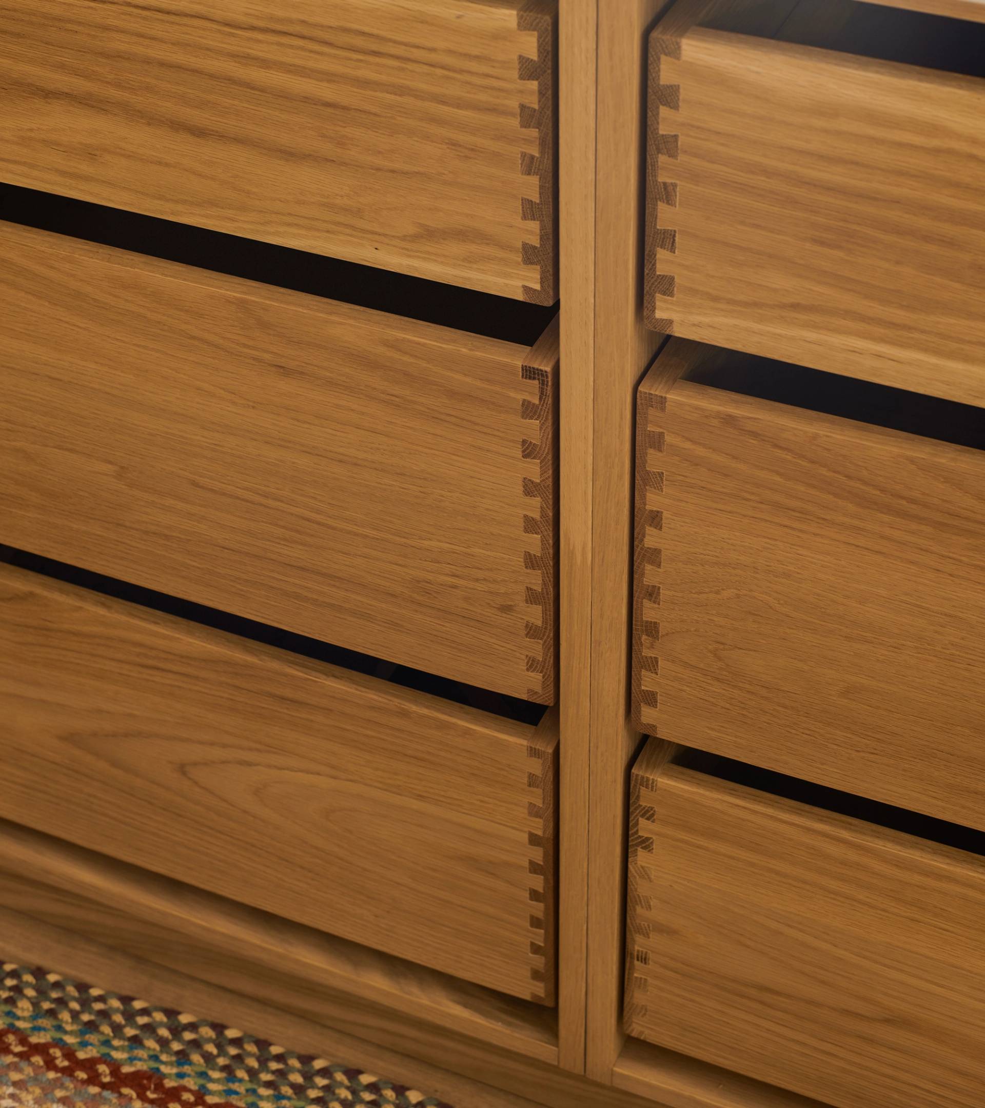 Close-up of a wooden kitchen cabinet with six visible drawers featuring dovetail joints. The cabinet has a natural wood finish and is topped with a light-colored countertop. A dark pot is partially visible on the counter.