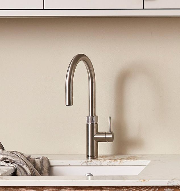 A modern stainless steel kitchen faucet is mounted on a marble countertop. A striped dish towel is draped to the left of the sink. The background features a light beige wall and white cabinetry.