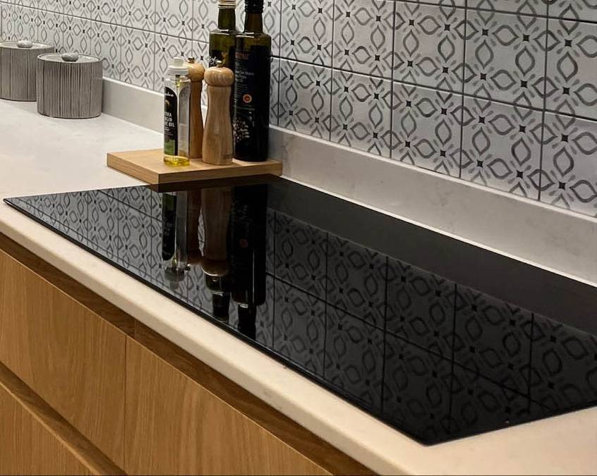 Modern kitchen counter featuring a black induction cooktop. Behind are bottles of olive oil and a wooden tray with salt and pepper shakers. The backsplash has a geometric tile pattern, and the cabinetry is light wood
