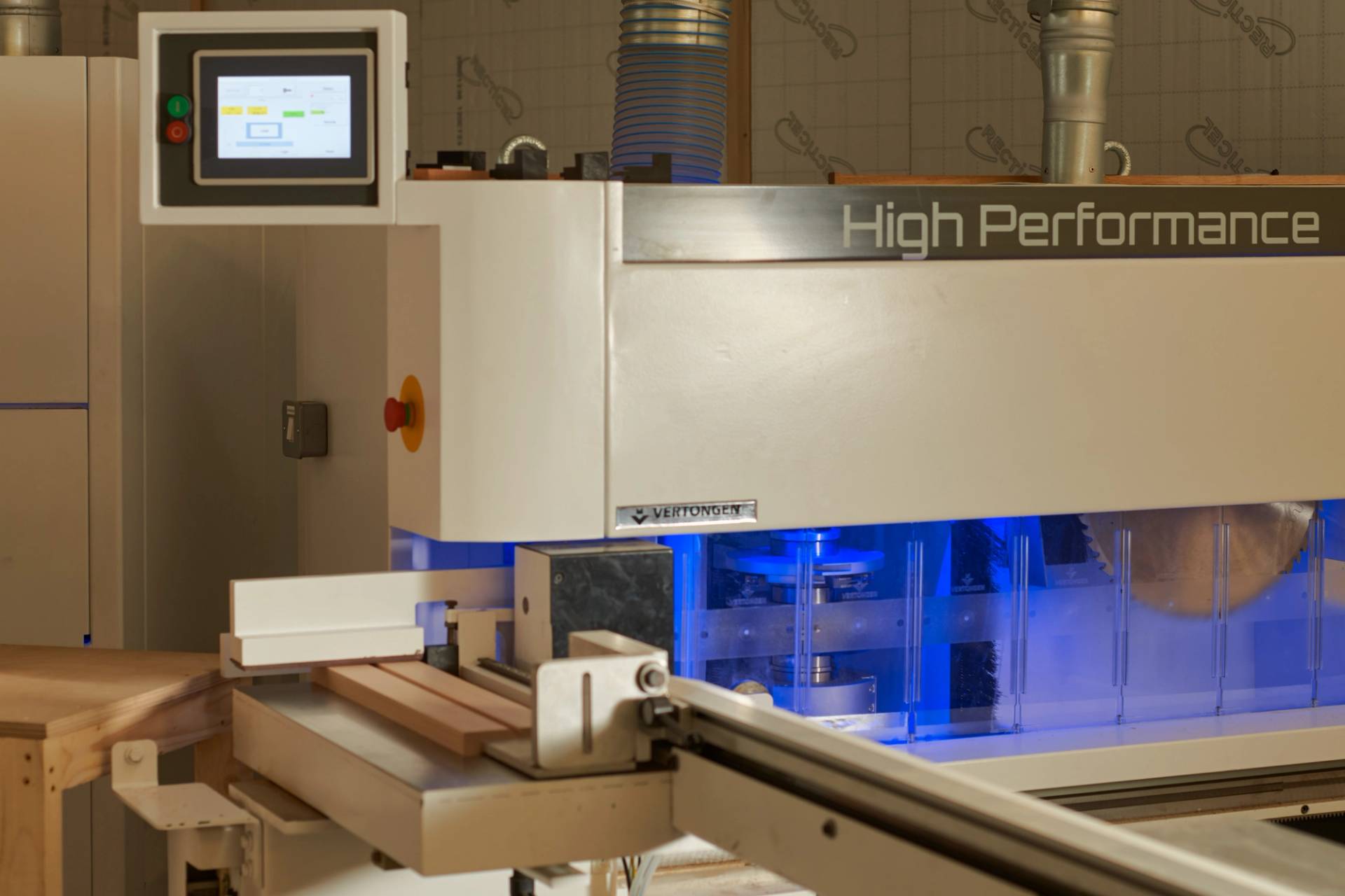 A high-performance industrial machine with a digital display and blue-lit inner components processes wooden boards. The surrounding area includes ductwork and a control panel.