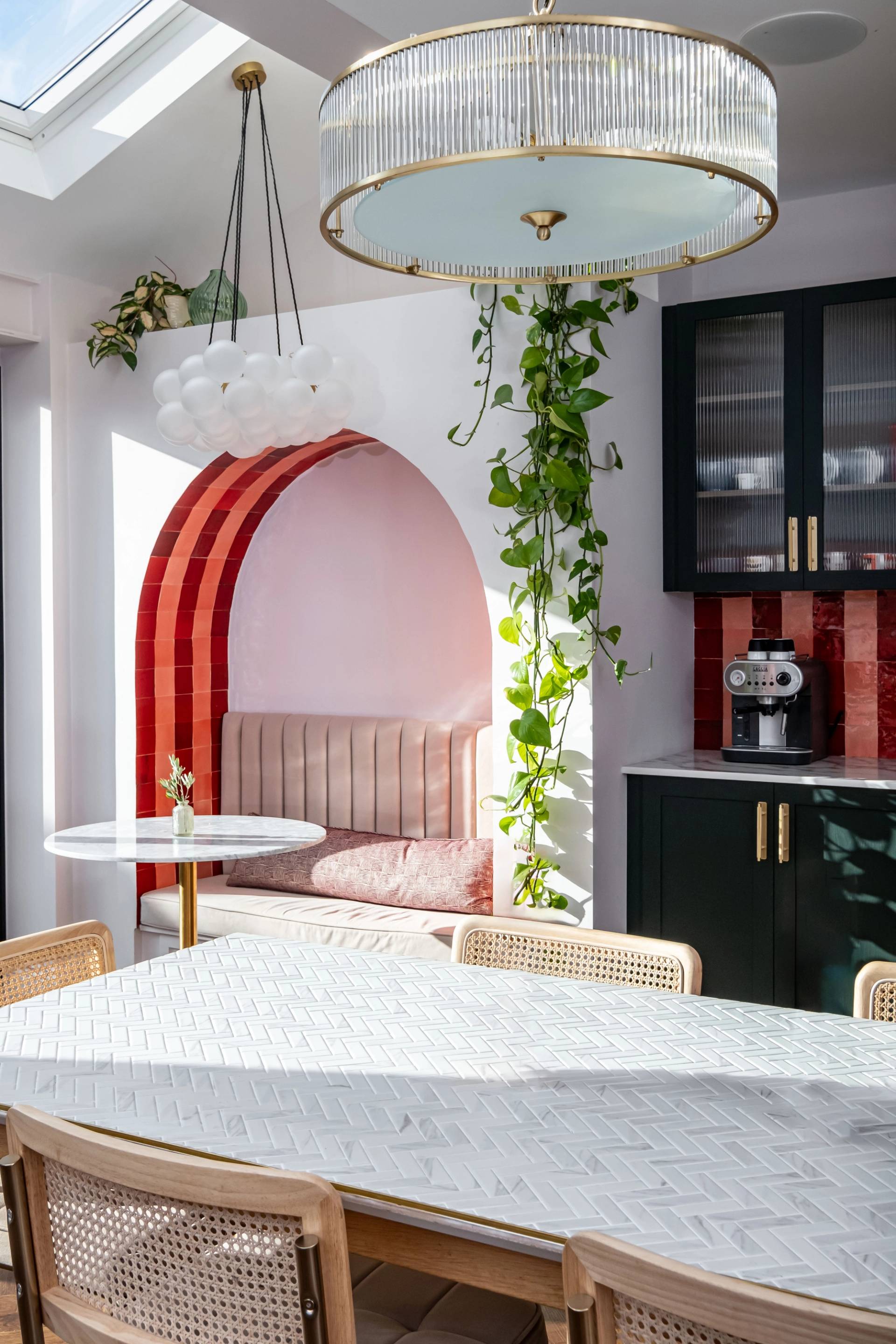 A bright, modern kitchen with a skylight. The room features a white dining table with candles, cane-back chairs, a cozy built-in bench with pink cushions under an archway, red tile backsplash, dark green cabinets, and hanging plants. Two elegant light fixtures hang from the ceiling.