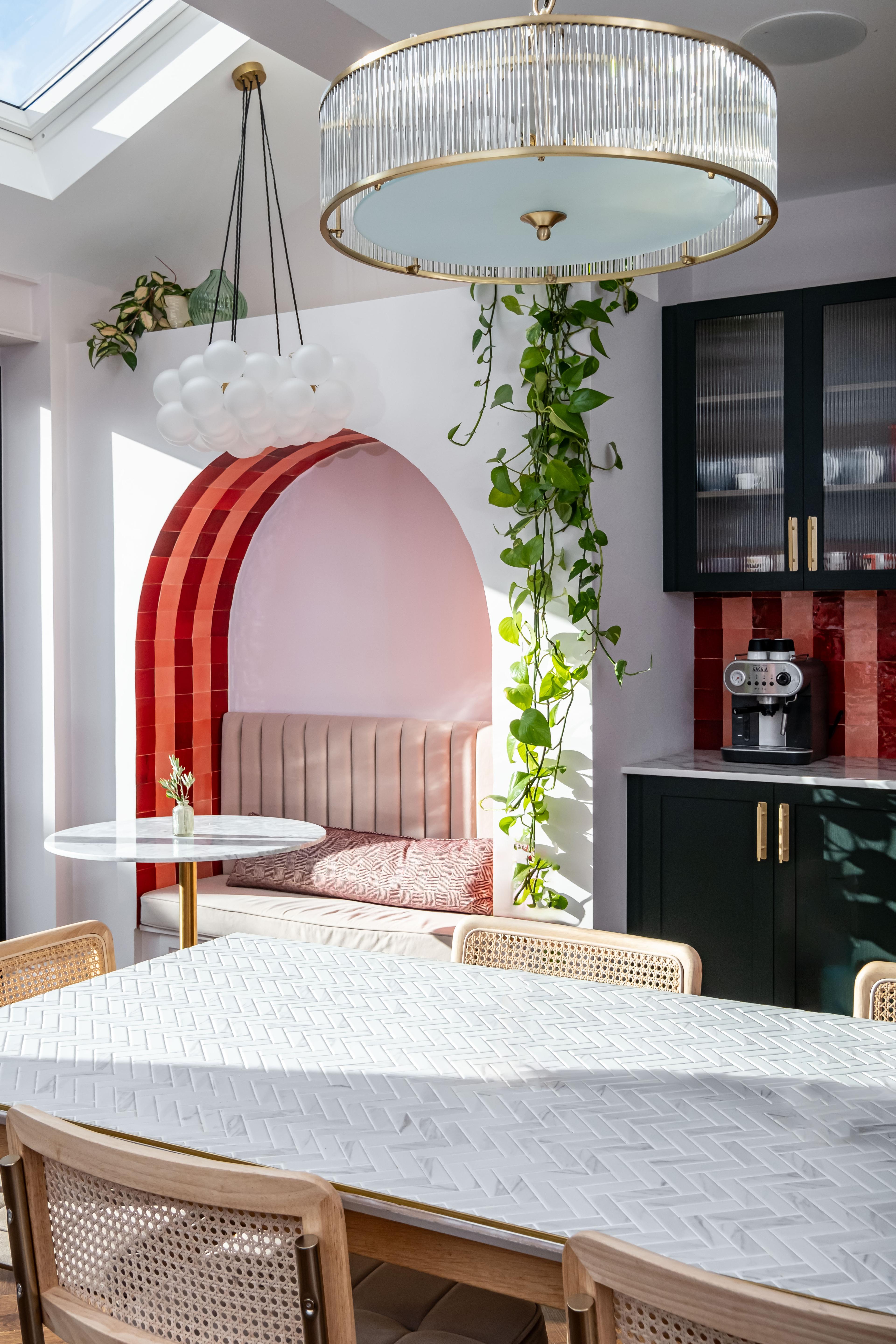 A bright, modern kitchen with a skylight. The room features a white dining table with candles, cane-back chairs, a cozy built-in bench with pink cushions under an archway, red tile backsplash, dark green cabinets, and hanging plants. Two elegant light fixtures hang from the ceiling.
