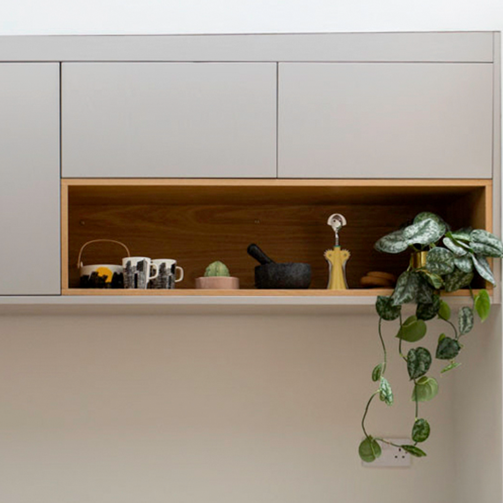 A modern kitchen shelf with mugs, a teapot, a mortar and pestle, a yellow pepper grinder, a small plant, and a trailing vine plant hanging down the side. The shelf is built into minimalist cabinetry.