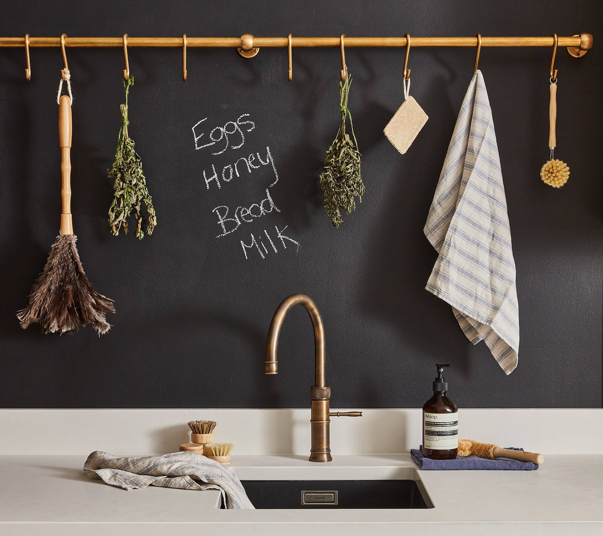 A kitchen sink scene with a bronze faucet. Above it, dried herbs, brushes, and a cloth hang on a golden rod. A blackboard wall lists Eggs, Honey, Bread, Milk. Nearby are a soap dispenser and a scrub brush on the counter.