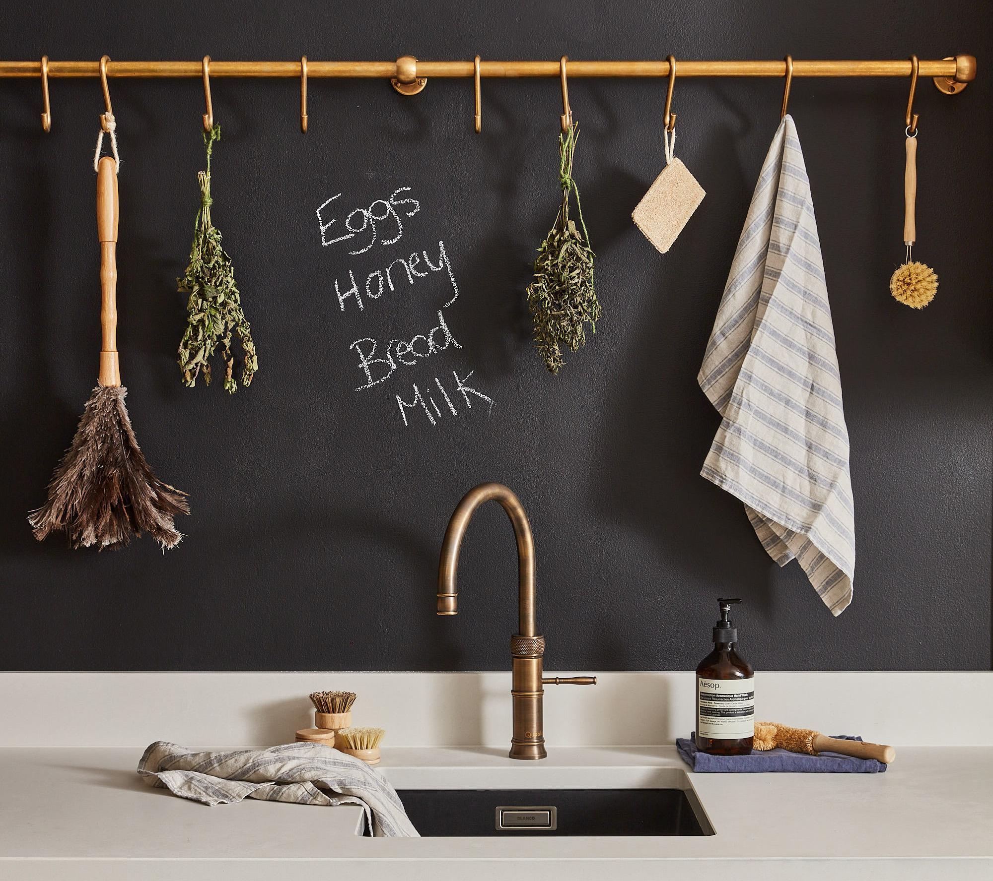 A kitchen sink scene with a bronze faucet. Above it, dried herbs, brushes, and a cloth hang on a golden rod. A blackboard wall lists Eggs, Honey, Bread, Milk. Nearby are a soap dispenser and a scrub brush on the counter.