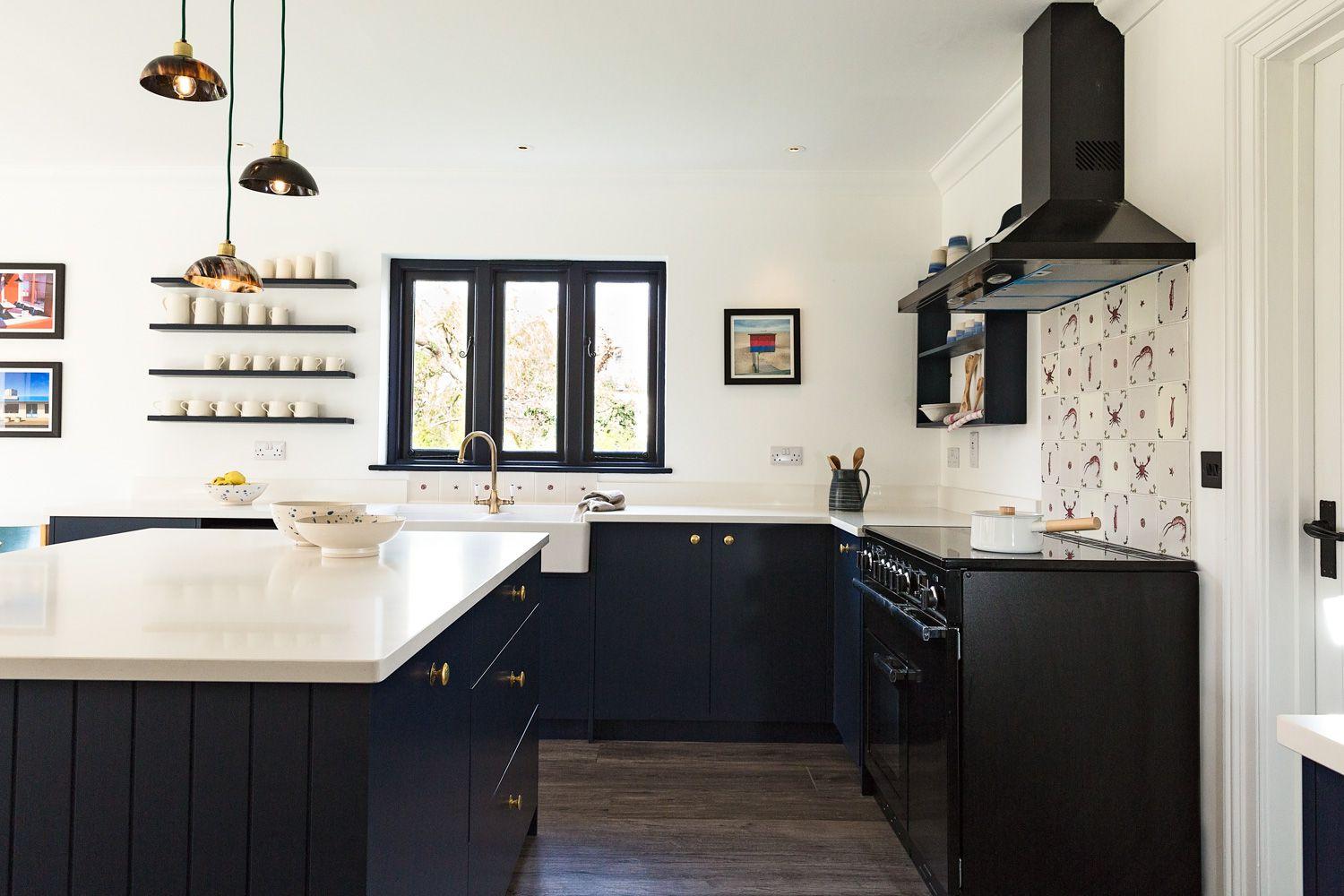 Modern kitchen with dark cabinets, white countertops, and a black stove. Pendant lights hang above a central island. Open shelves display dishes, and a picture window provides natural light. Decorative backsplash adds a touch of color.