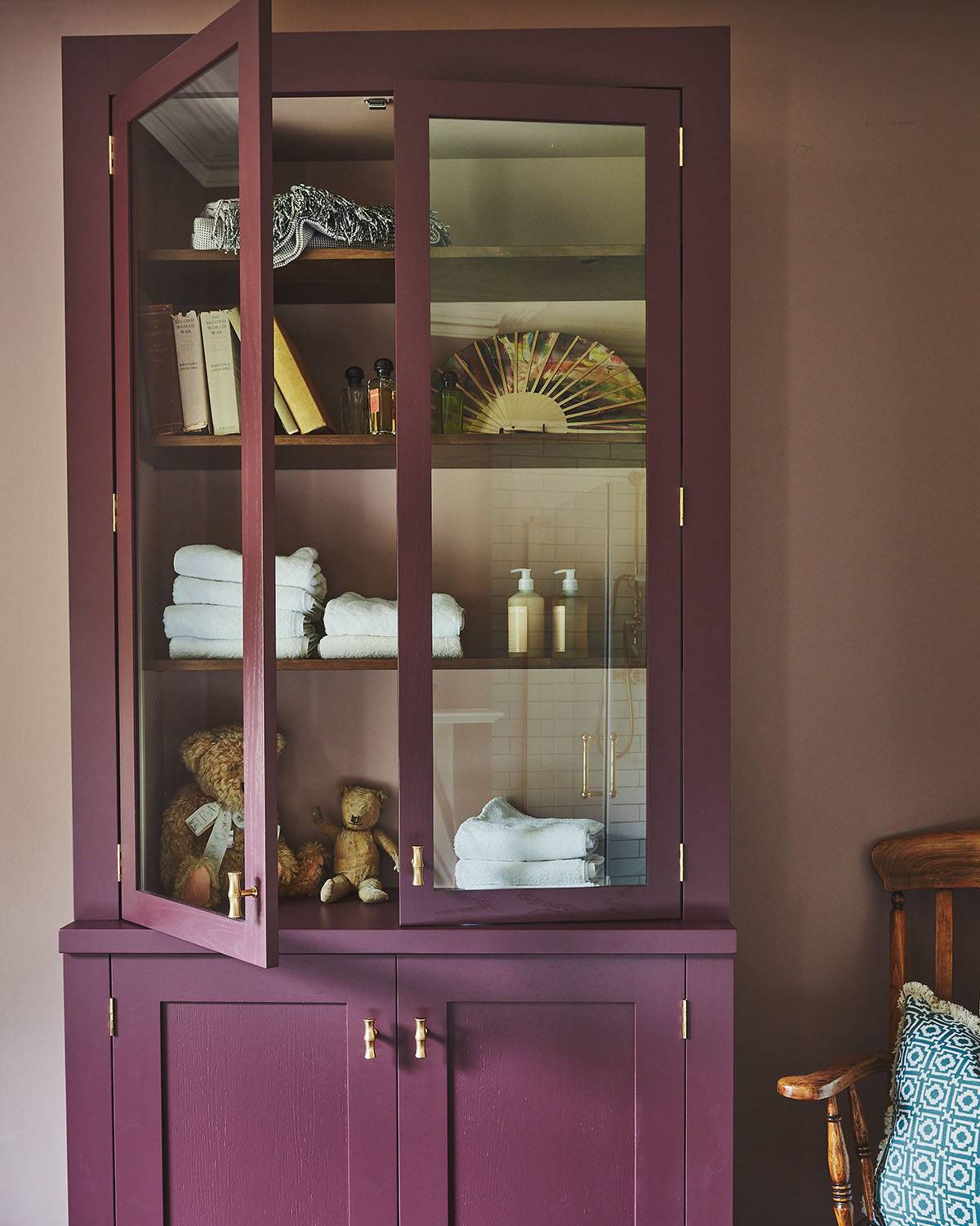 A bathroom with mauve walls features a tall, deep purple cabinet with glass doors displaying towels, books, and a teddy bear. A toilet, white bathtub, and wooden chair with a patterned cushion are also visible.