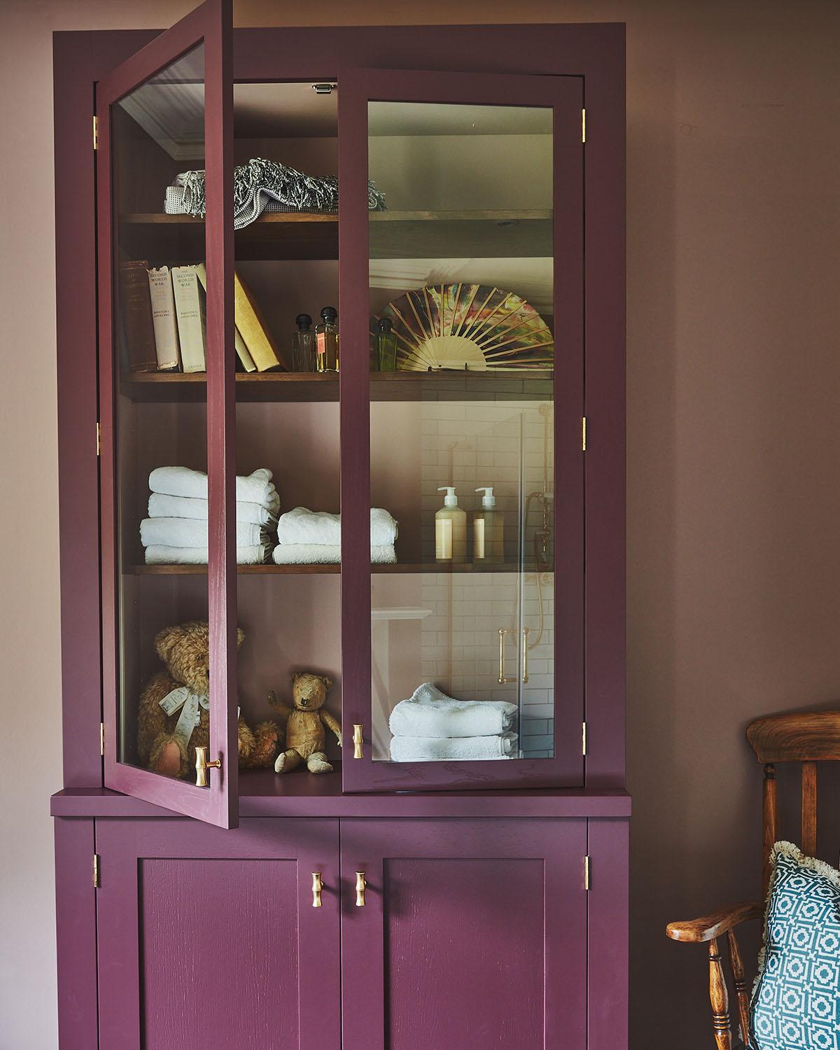 A bathroom with mauve walls features a tall, deep purple cabinet with glass doors displaying towels, books, and a teddy bear. A toilet, white bathtub, and wooden chair with a patterned cushion are also visible.
