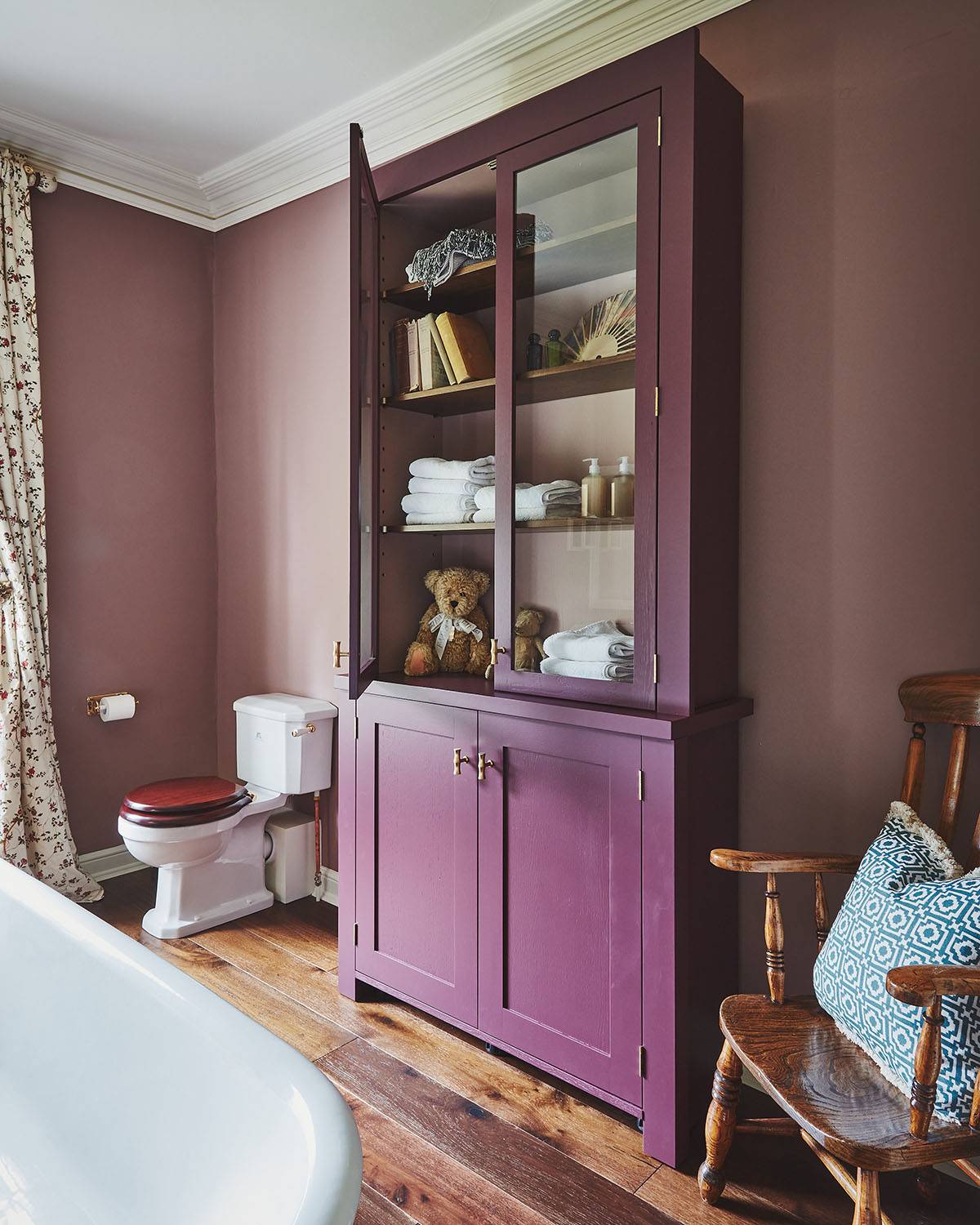 A bathroom with mauve walls features a tall, deep purple cabinet with glass doors displaying towels, books, and a teddy bear. A toilet, white bathtub, and wooden chair with a patterned cushion are also visible.