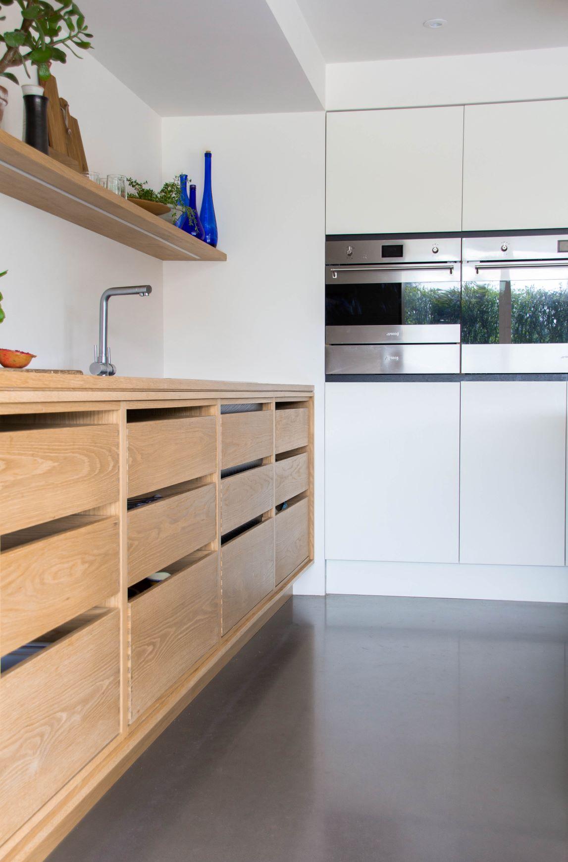 Modern kitchen with light wood cabinets and drawers, a stainless steel faucet, and a smooth countertop. The background has a white wall with built-in stainless steel appliances. Decorative plants and blue bottles adorn the open shelf.