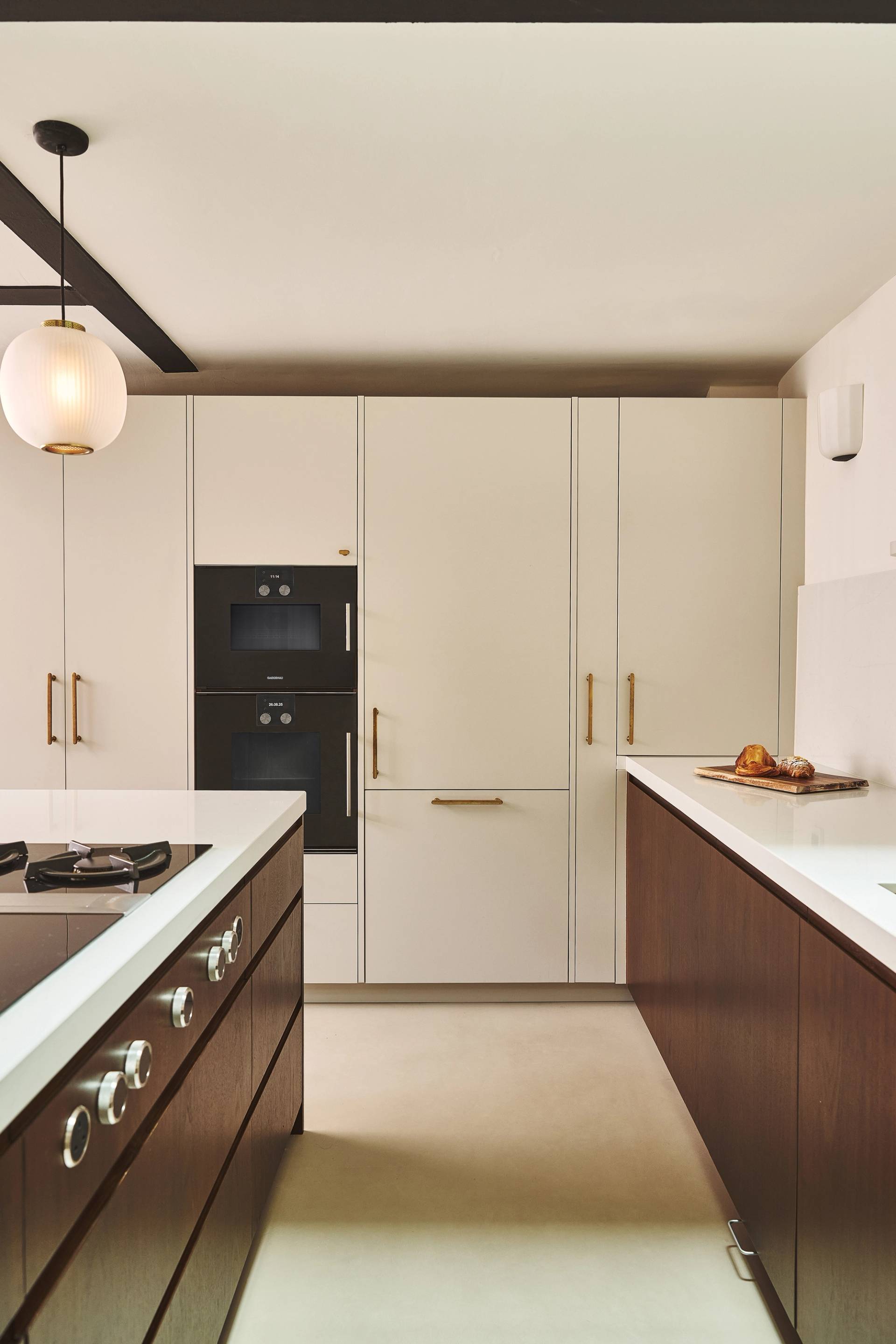 Modern kitchen with cream-colored cabinets, gold handles, dark wood lower cabinets, built-in black oven, white countertops, and a round hanging light fixture. A loaf of bread sits on the counter.
