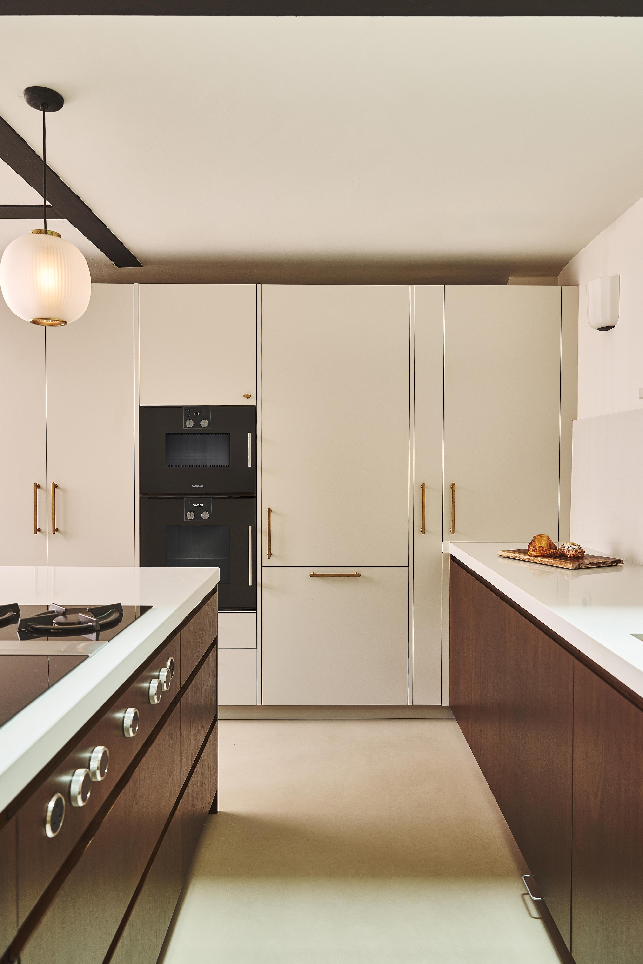 Modern kitchen with cream-colored cabinets, gold handles, dark wood lower cabinets, built-in black oven, white countertops, and a round hanging light fixture. A loaf of bread sits on the counter.