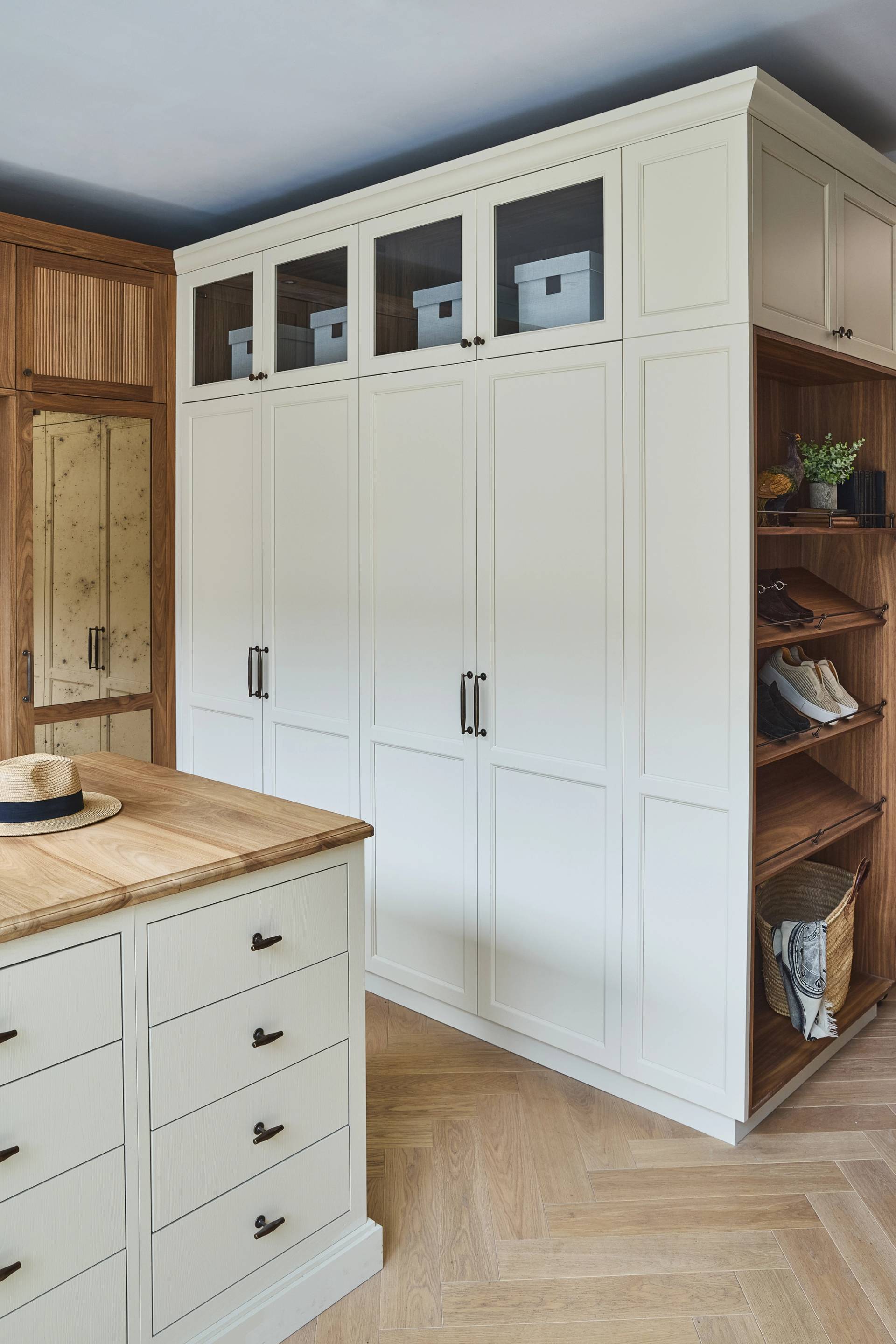 Modern dressing room with tall white cabinetry and walnut internals. Island chest of drawers in foreground with straw hat placed on top. Wardrobes in background with shoe rack shelving on the right side.