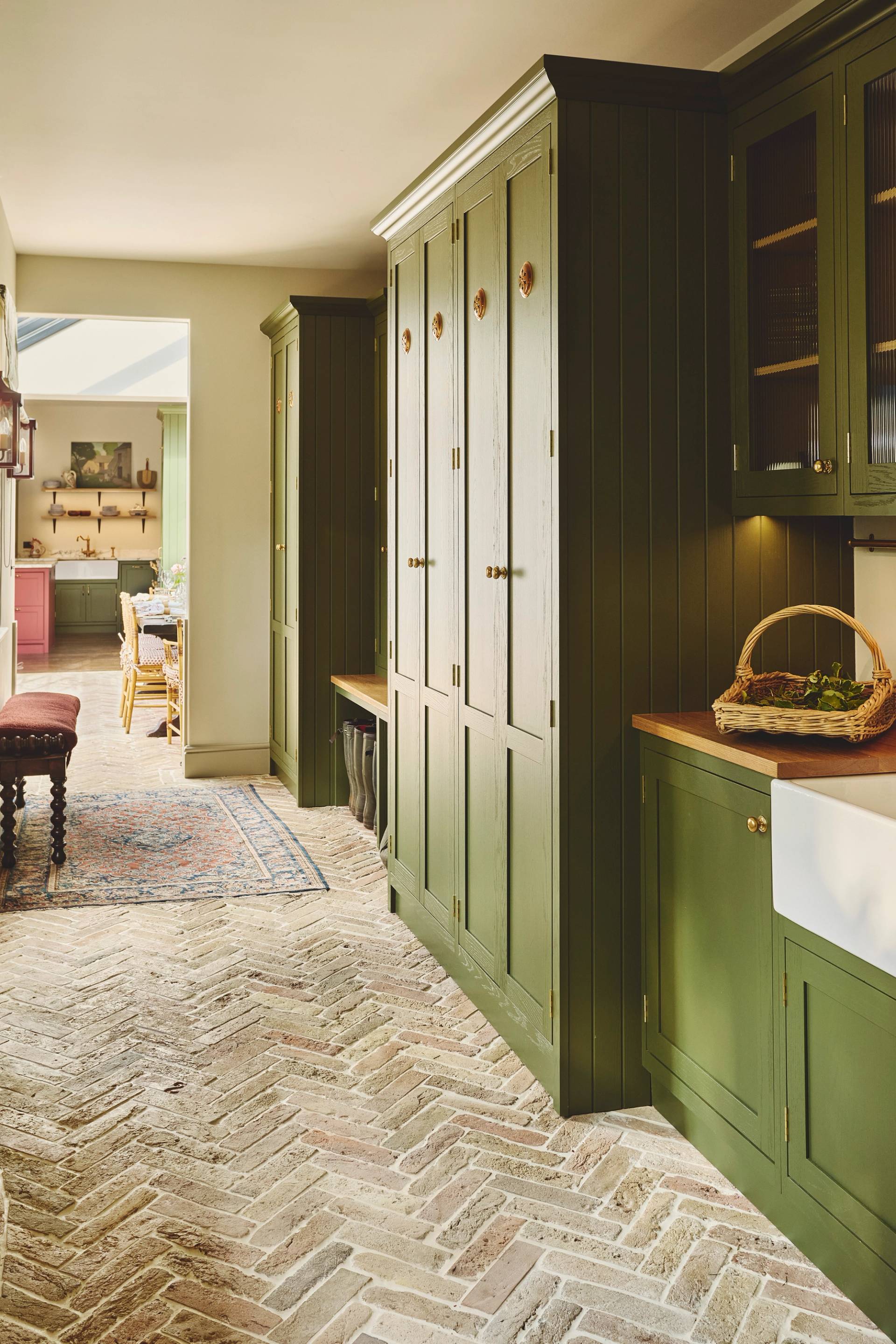 A laundry area with olive green cabinets, a stacked washer and dryer inside a closet, woven basket above, a farmhouse sink, and herringbone patterned tile floor.