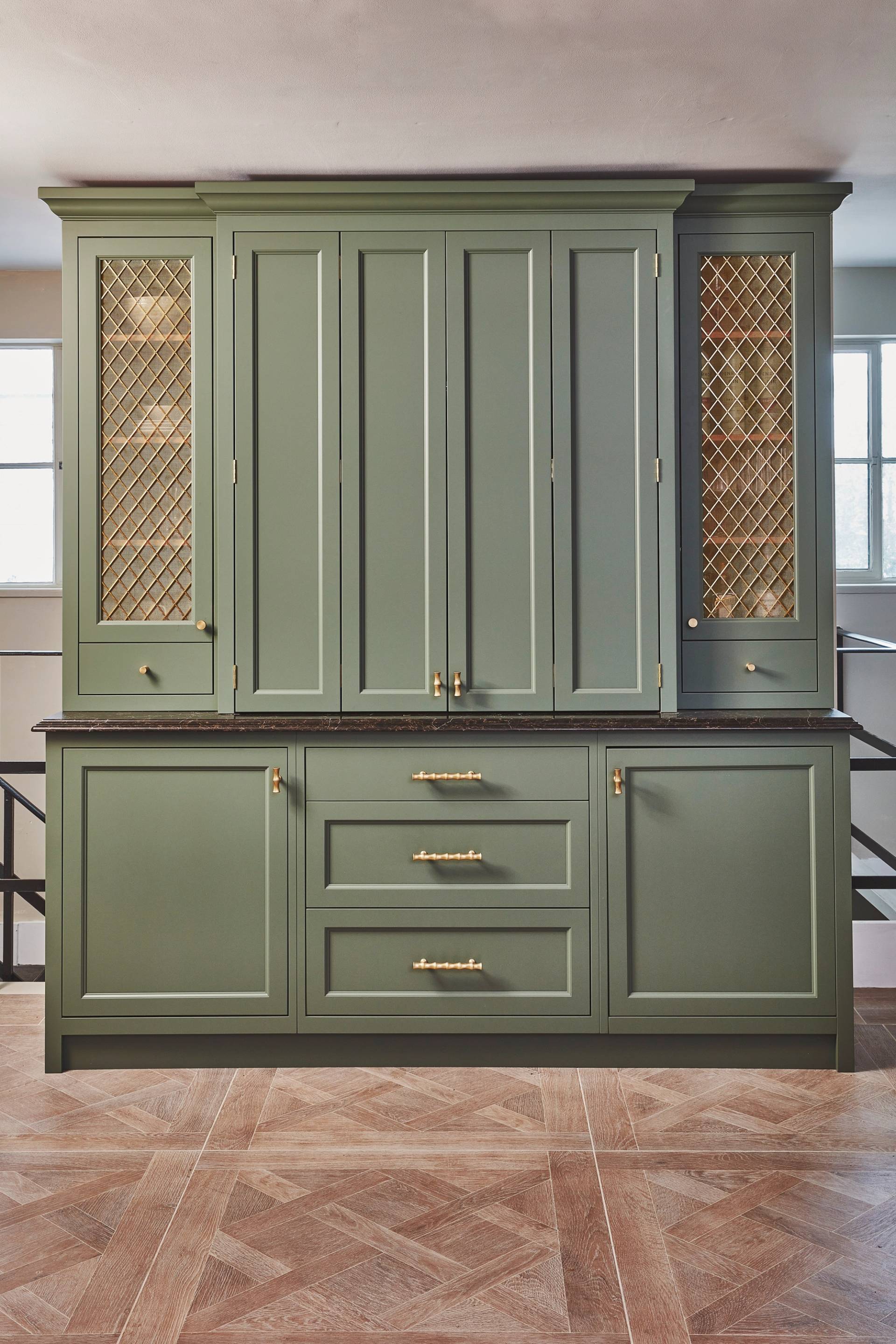 Green kitchen cabinets with gold handles and decorative gold mesh panels on the upper side sections, set against a neutral wall and patterned wood floor.