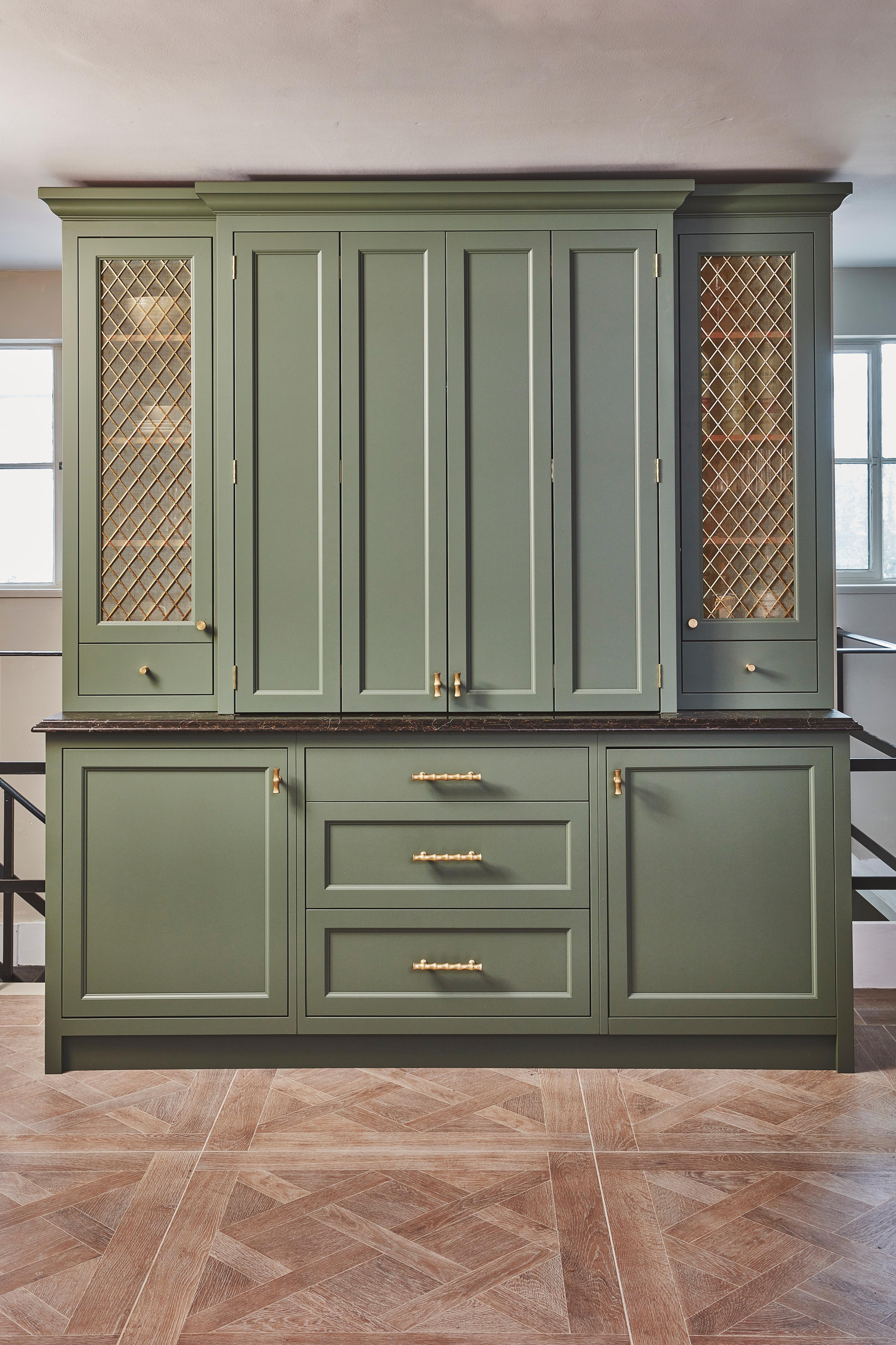 Green kitchen cabinets with gold handles and decorative gold mesh panels on the upper side sections, set against a neutral wall and patterned wood floor.
