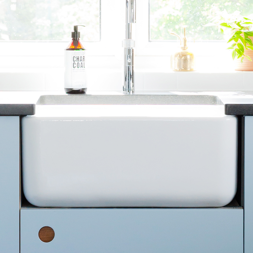 A bright kitchen scene with a white farmhouse sink, a faucet, a bottle labeled Charcoal on the counter, a soap dispenser, and a green potted plant by a window with sunlight streaming in.