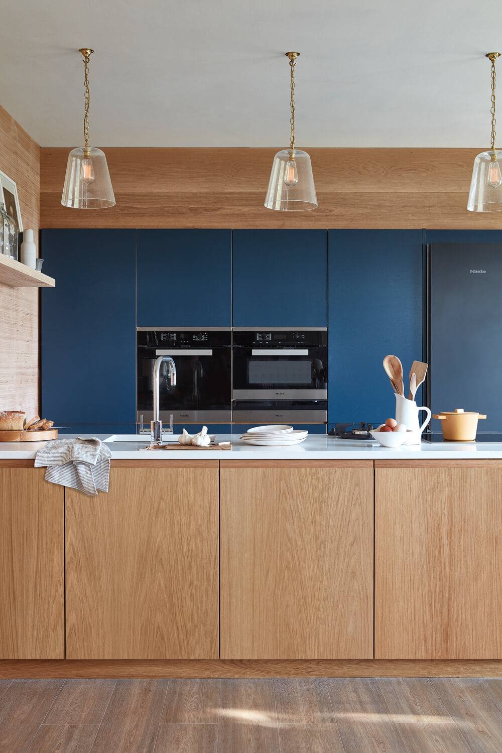 Modern kitchen with wood cabinets and blue accent paneling. Features a sleek countertop with kitchenware, built-in oven, and refrigerator. Three pendant lights hang from the ceiling, enhancing the warm, inviting atmosphere.