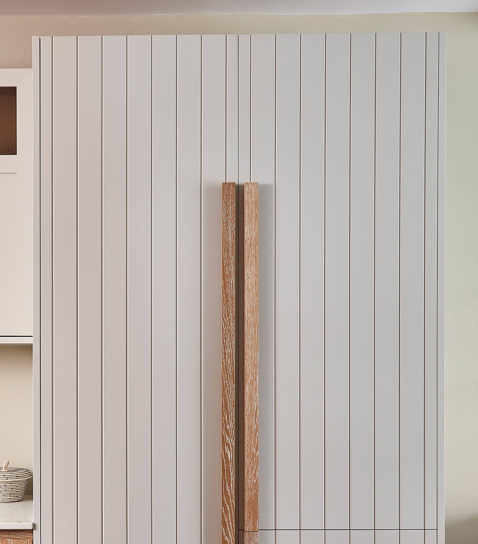 A minimalist kitchen setup featuring white paneled cabinetry with vertical grooves. The cabinet doors have long, slim, wooden handles in the center. A partial view of a shelf with a basket and jar is on the left.