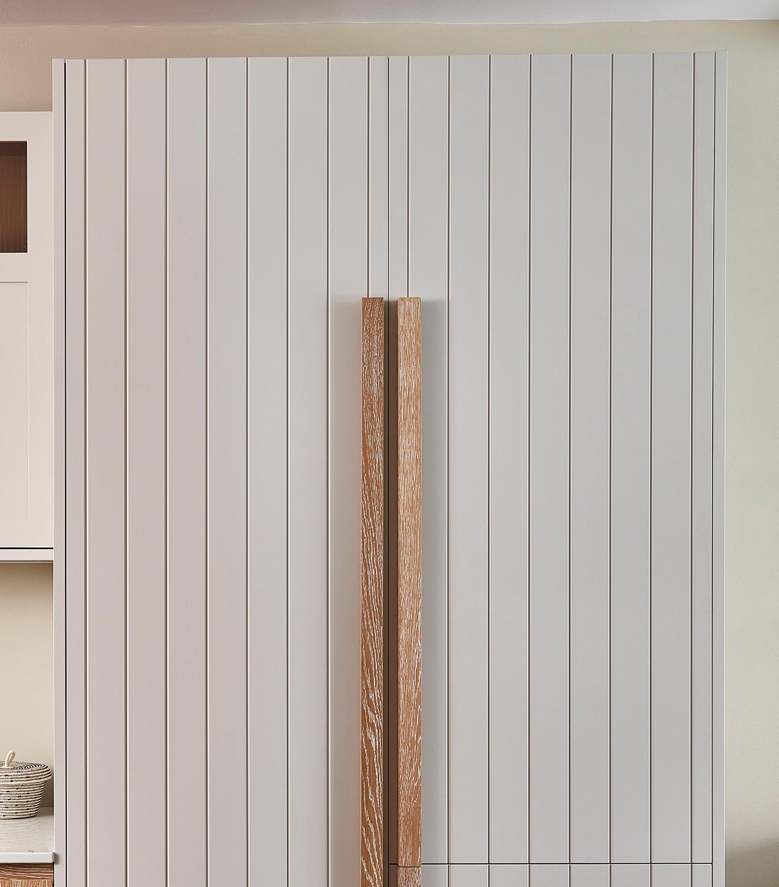 A minimalist kitchen setup featuring white paneled cabinetry with vertical grooves. The cabinet doors have long, slim, wooden handles in the center. A partial view of a shelf with a basket and jar is on the left.