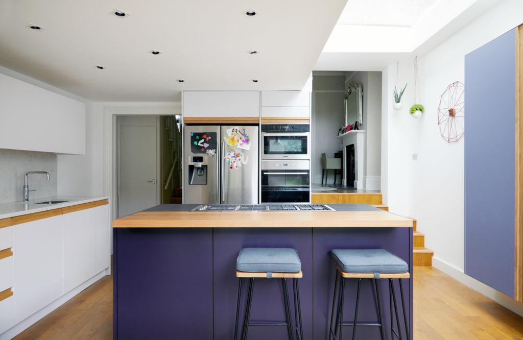 Modern open-plan kitchen with a deep purple central island, wood-topped counter with integrated gas hob, cushioned bar stools, and white surrounding cabinetry.