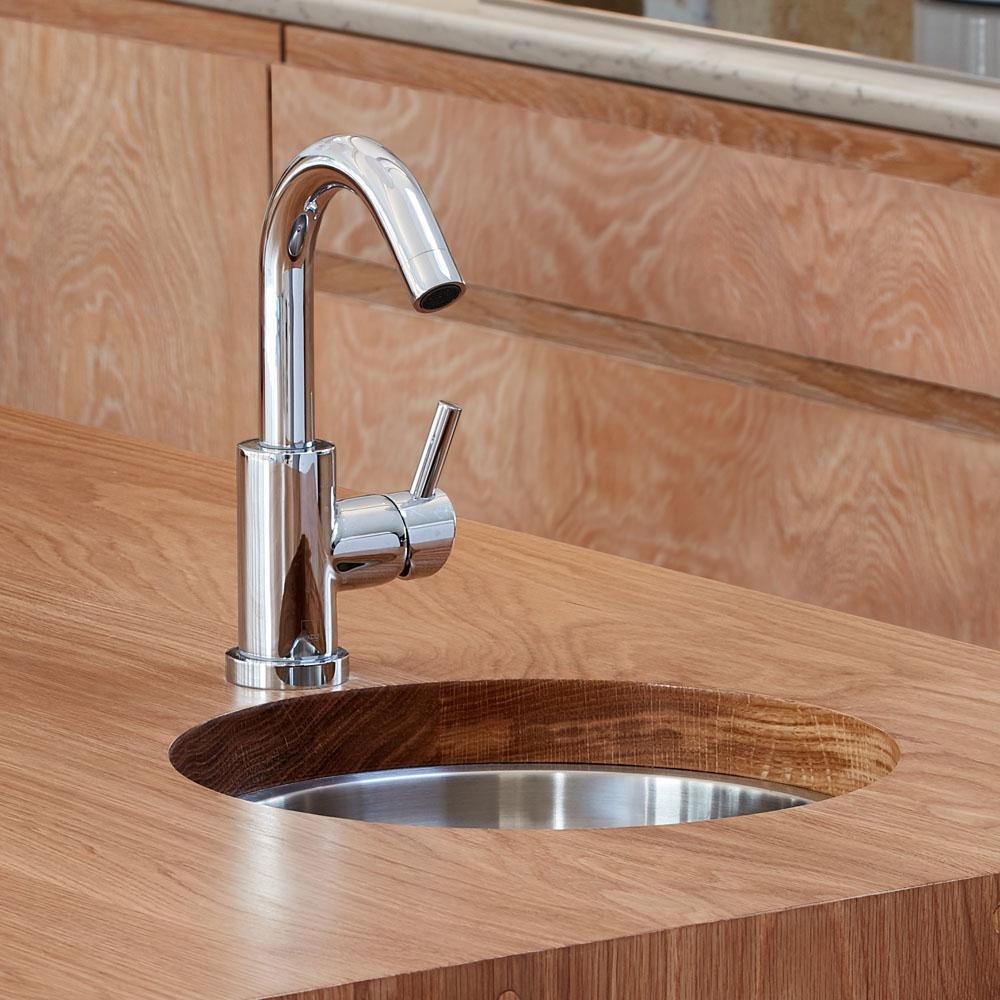 A modern kitchen faucet with a sleek, curved design is installed in a wooden countertop with a stainless steel sink. The faucet has a single lever handle for easy water control. The background features light wood cabinetry.