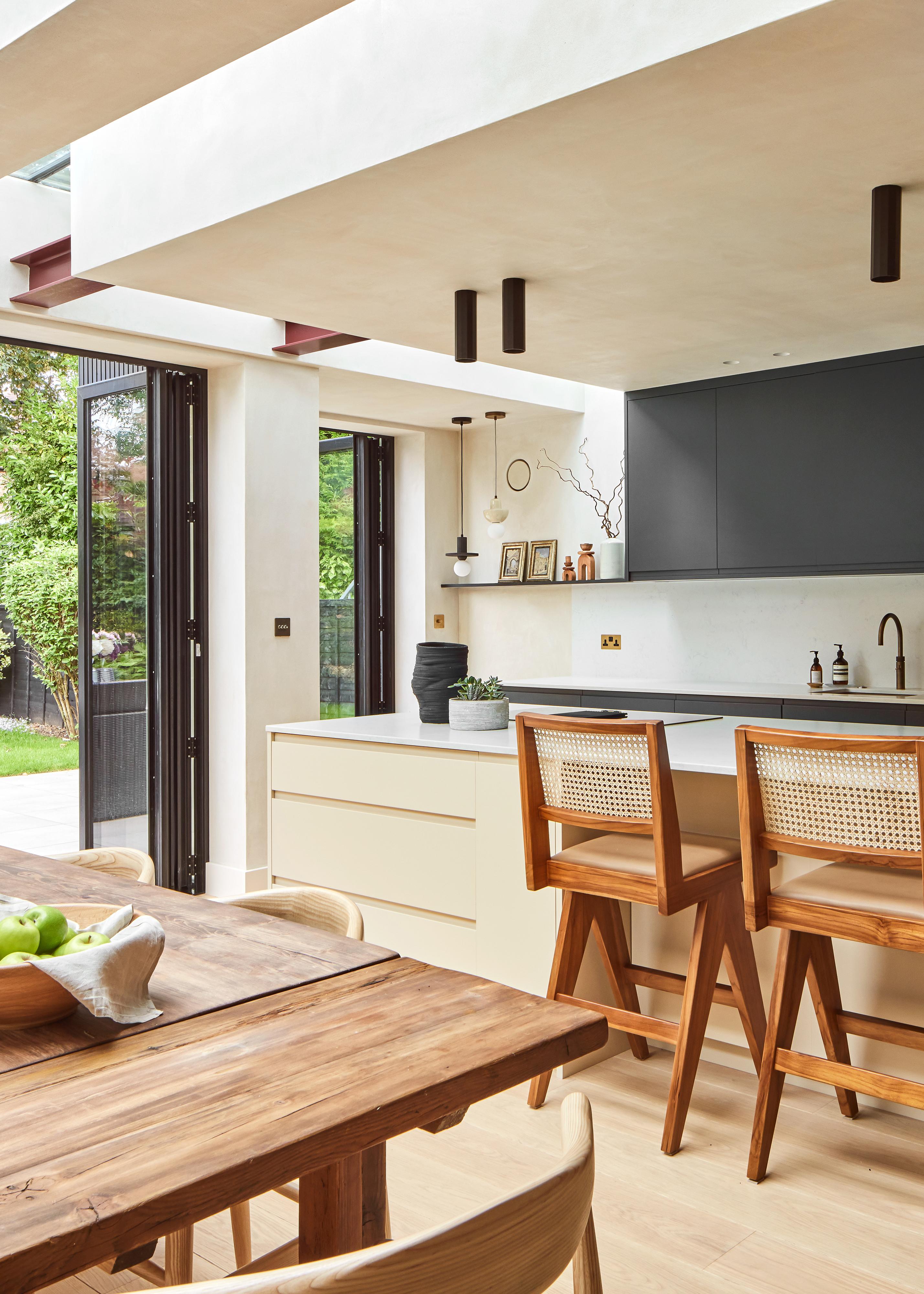Modern kitchen with an island, wooden bar stools, light cabinets, a wooden dining table, and large glass doors opening to a green garden. The space is bright and airy with minimalist decor.